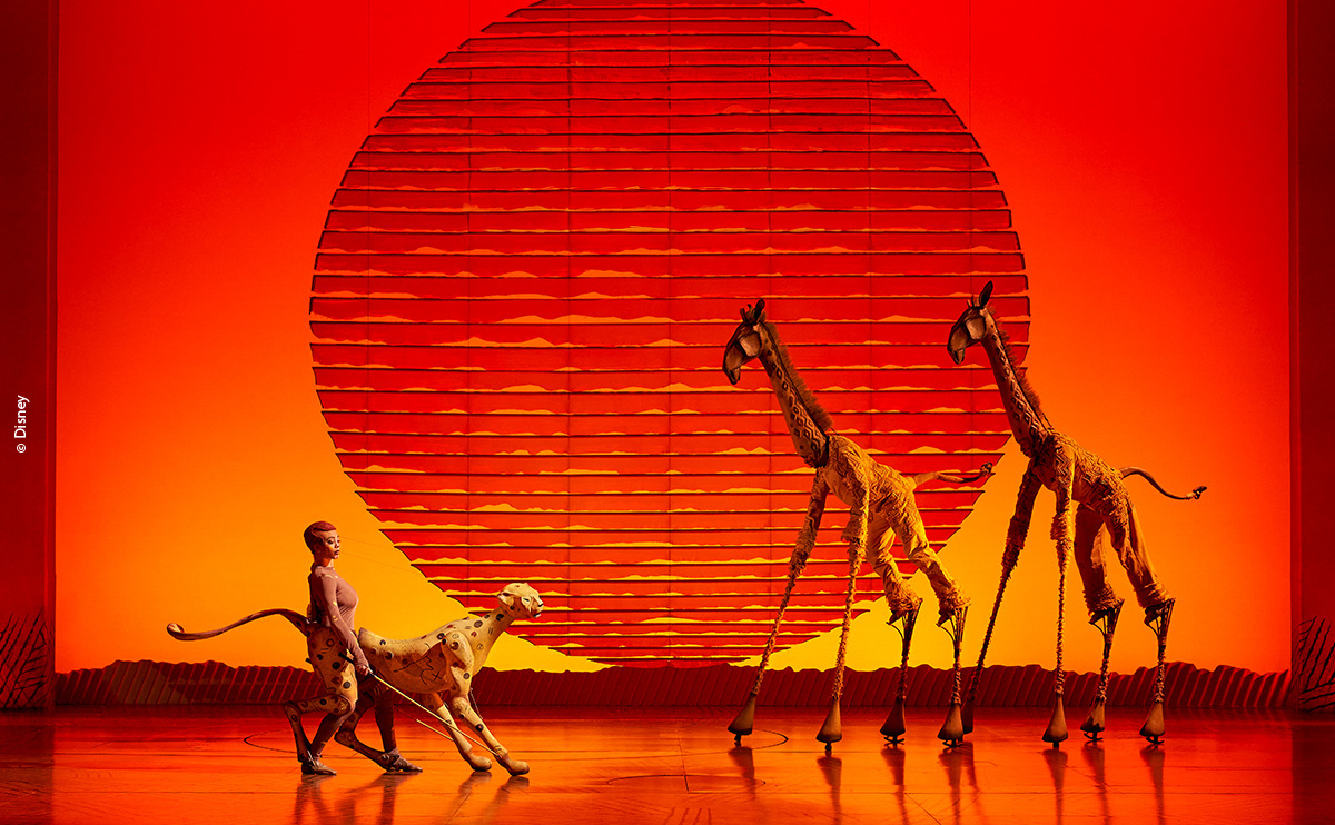 The Lion King musical stage with giraffe puppets and cheetah costume, red sunset.