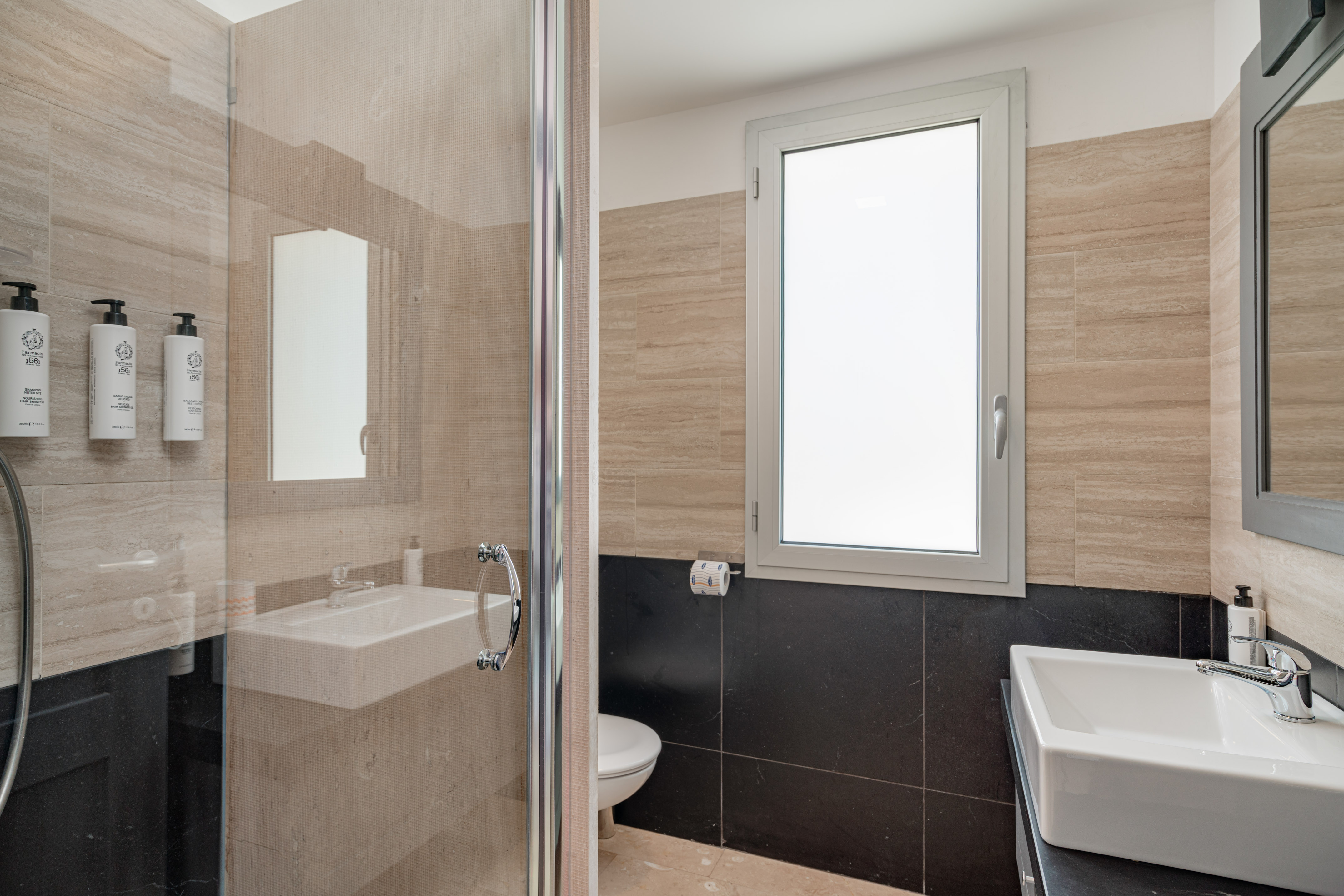 Modern bathroom with glass shower, two sinks, toilet, and light/dark tiled walls.
