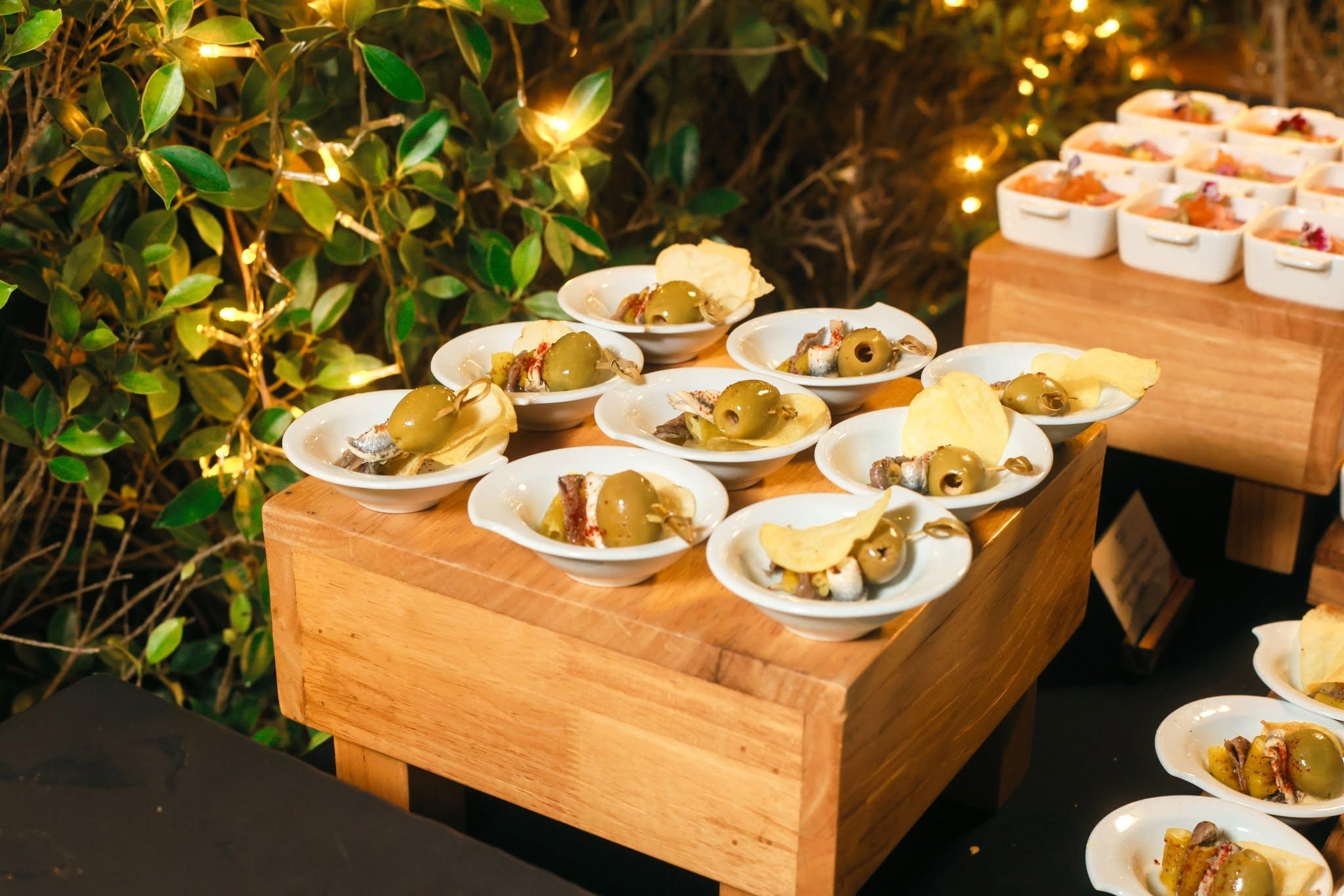 Catering display of tapas appetizers with olives, anchovies, and chips on wooden stands.