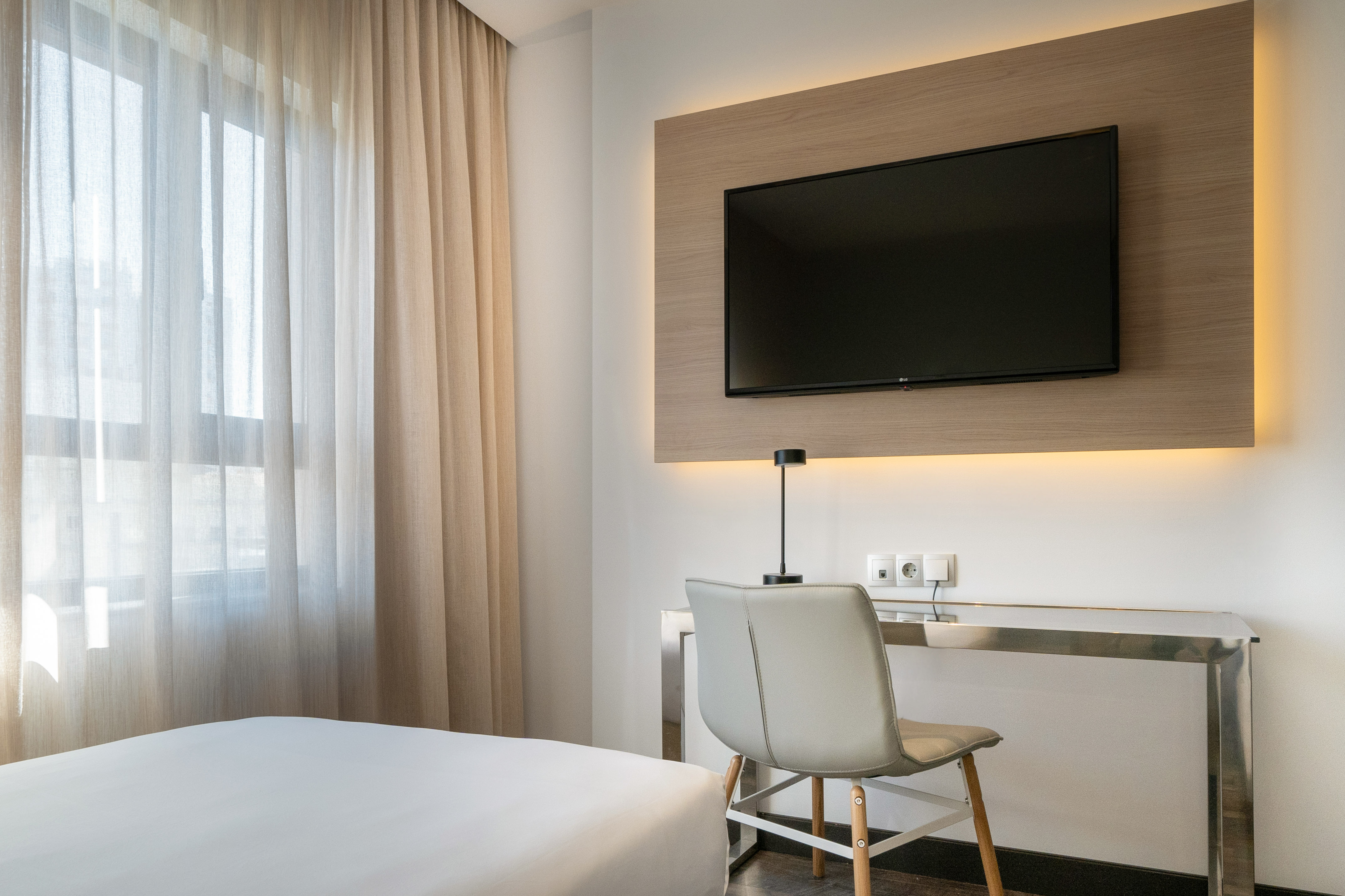 Modern hotel bedroom with wall-mounted TV, desk, and sheer window curtains.
