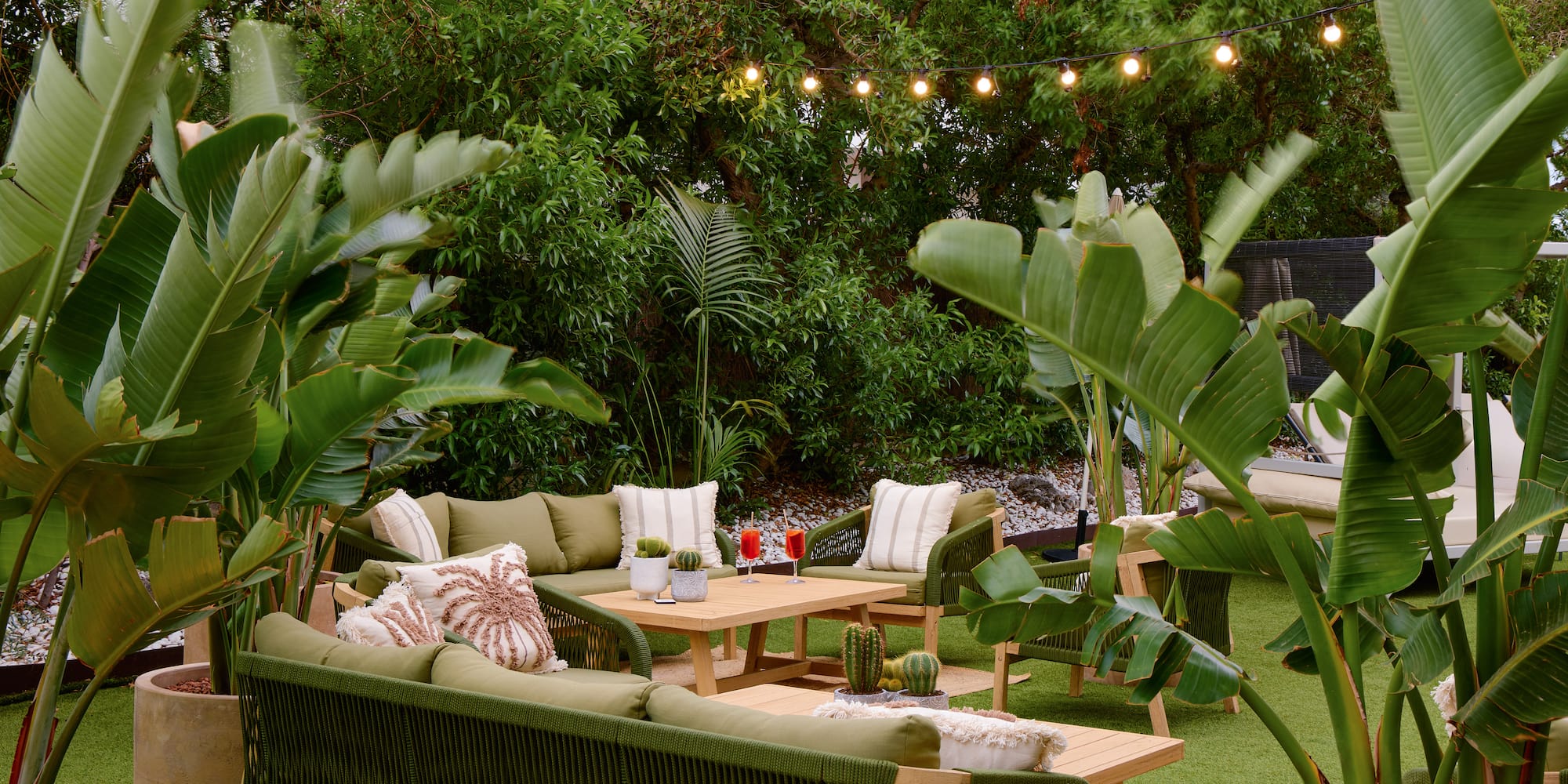 Tropical outdoor lounge with green patio furniture, lush plants, and string lights.