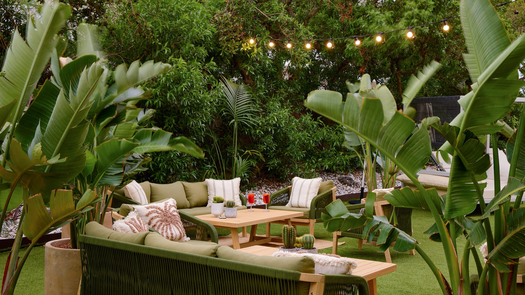 Tropical outdoor lounge with green patio furniture, lush plants, and string lights.