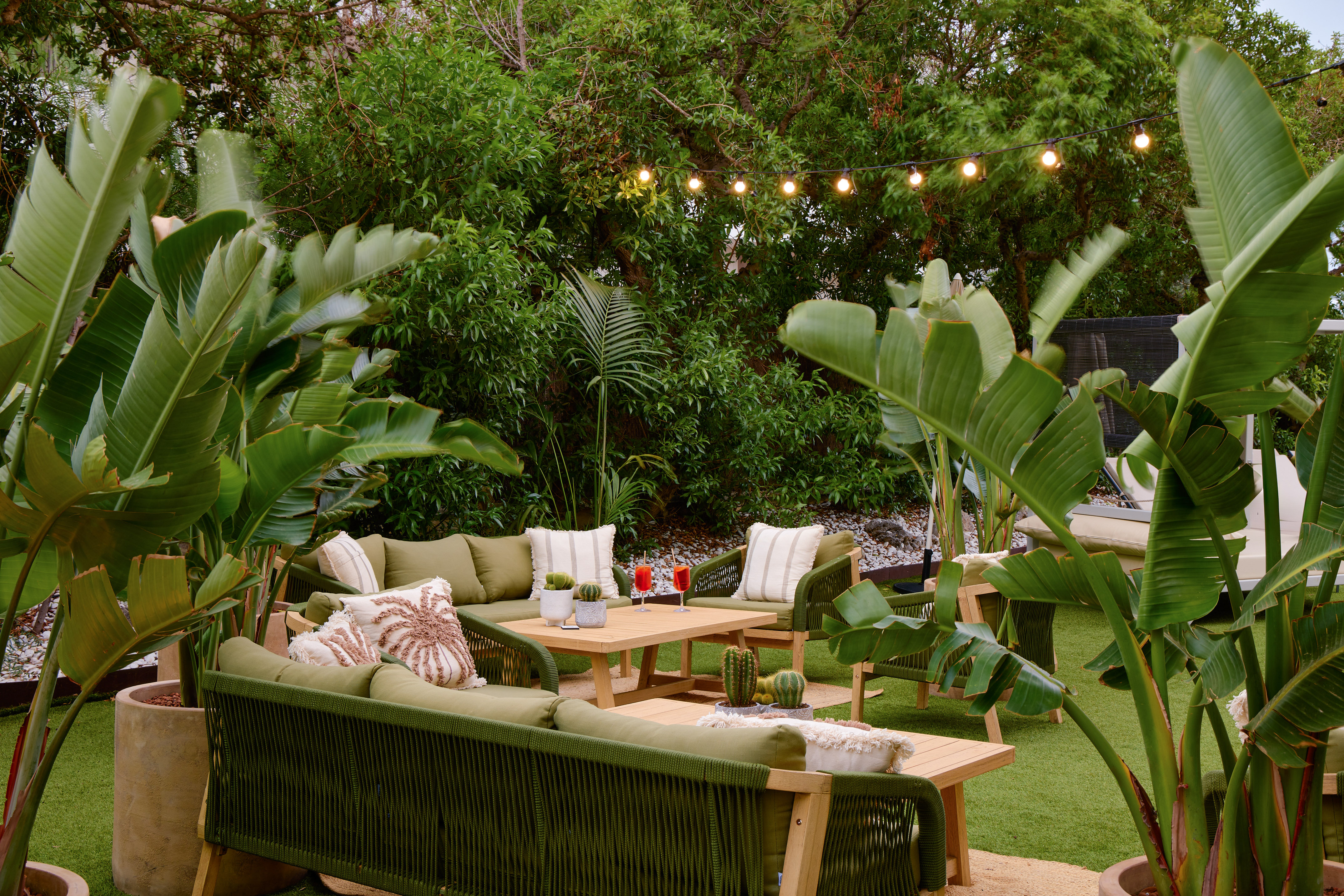 Tropical outdoor lounge with green patio furniture, lush plants, and string lights.