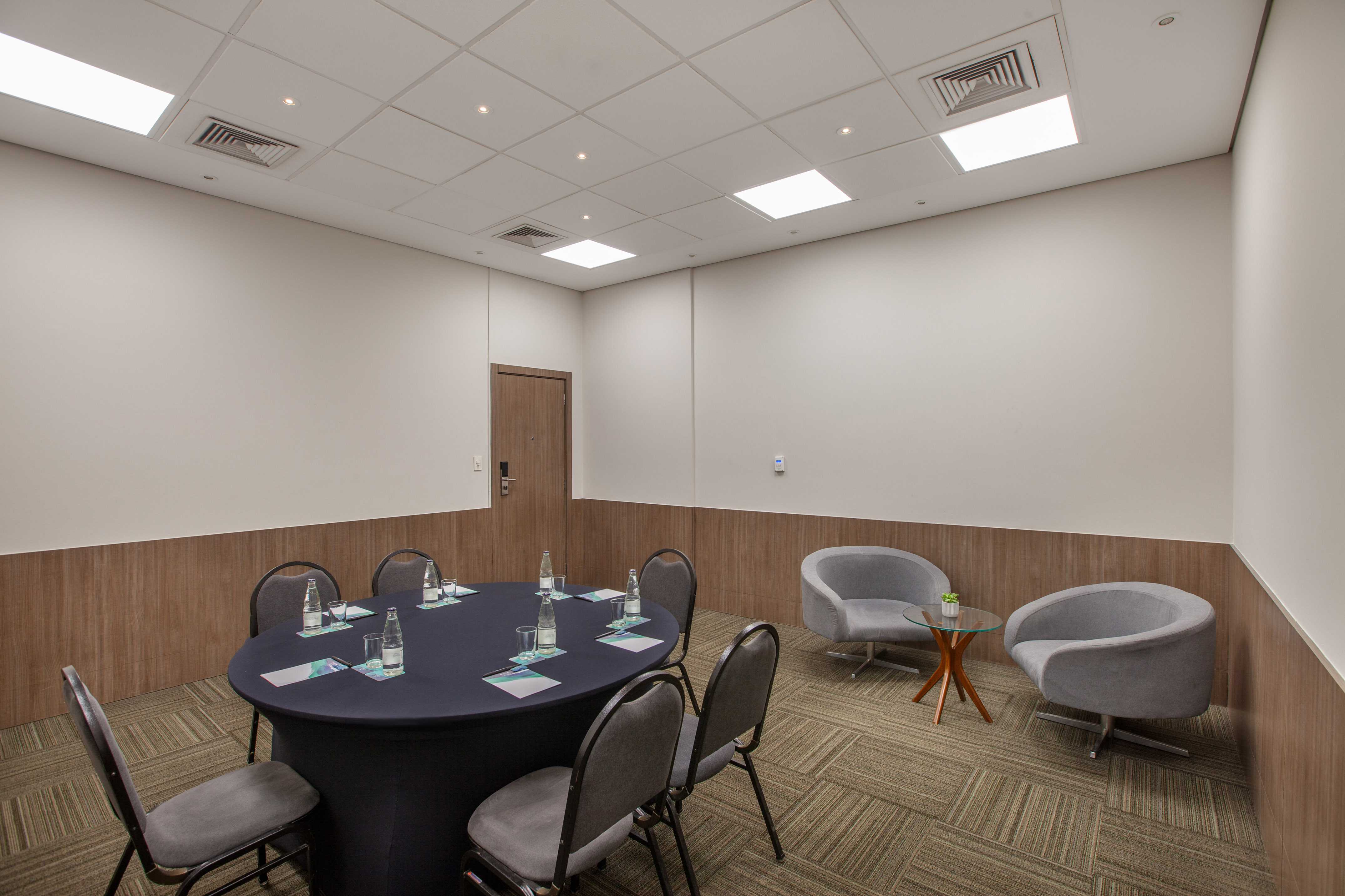 Modern small meeting room with round table, grey chairs, and a cozy lounge area.
