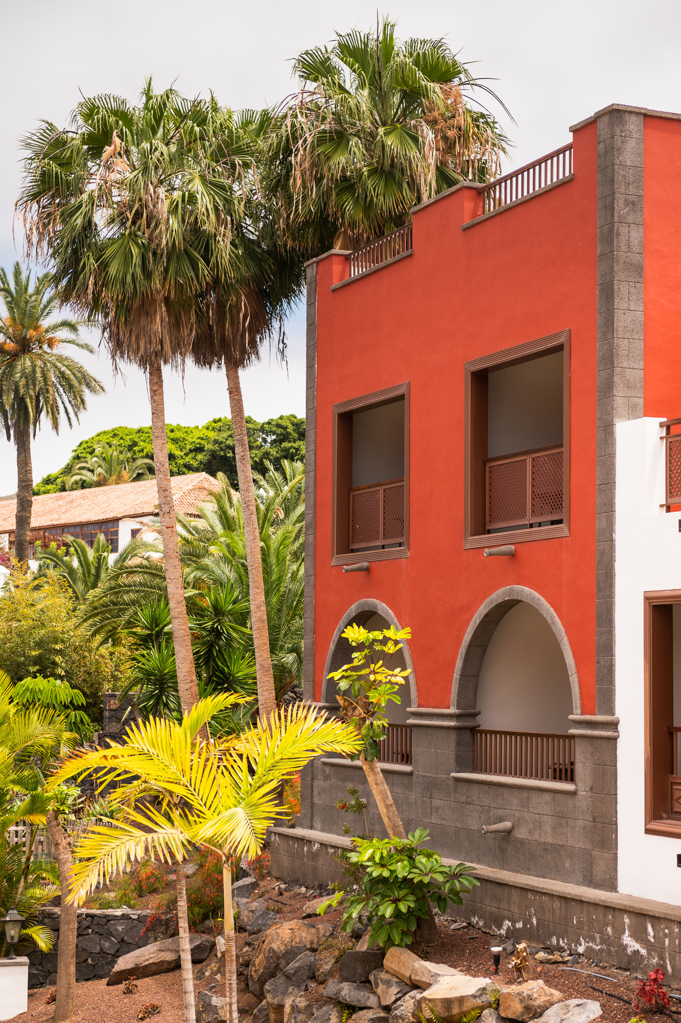 Red building with stone arches, balconies, and lush tropical palm trees.