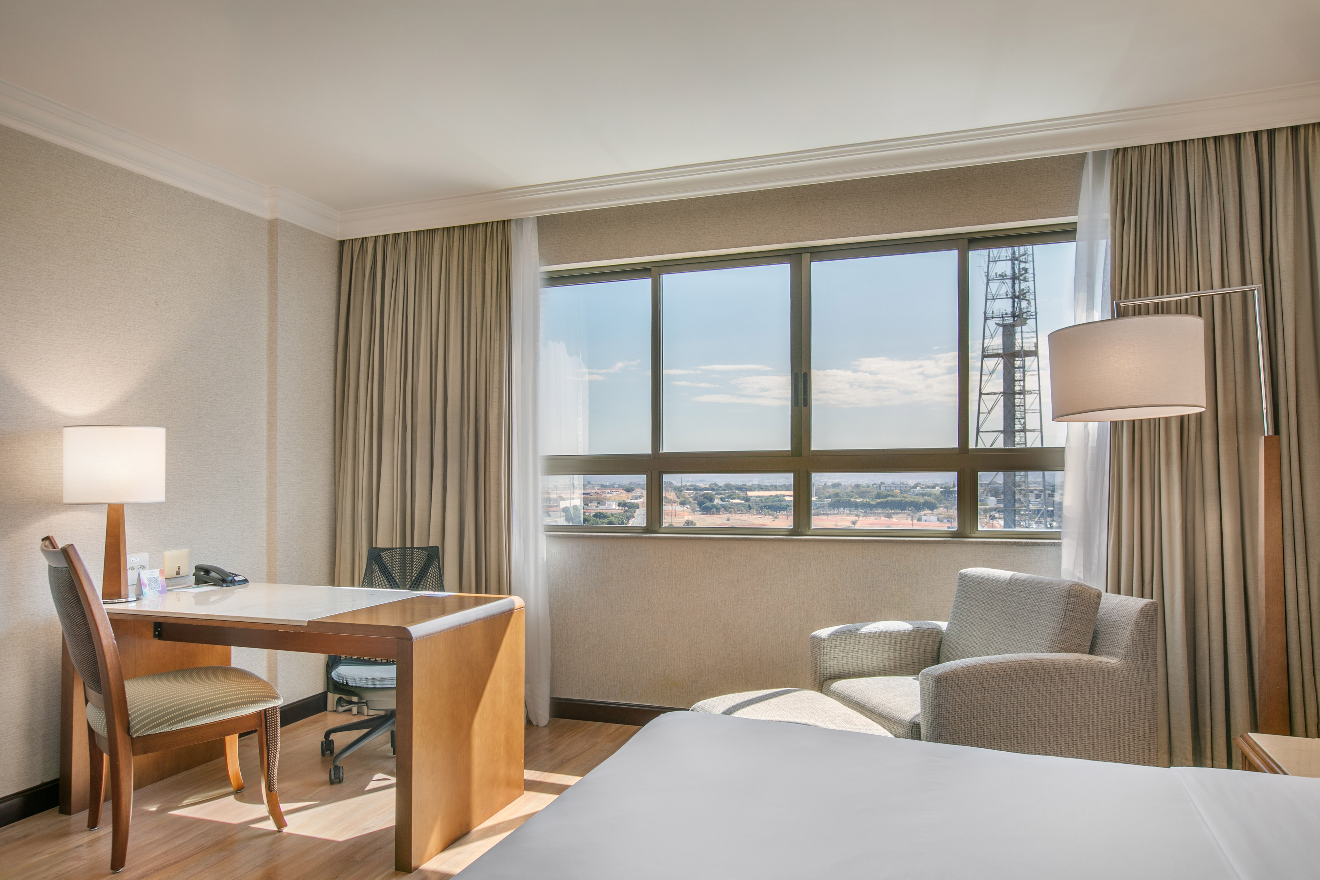 Hotel room with work desk, armchair, and window offering a city view.