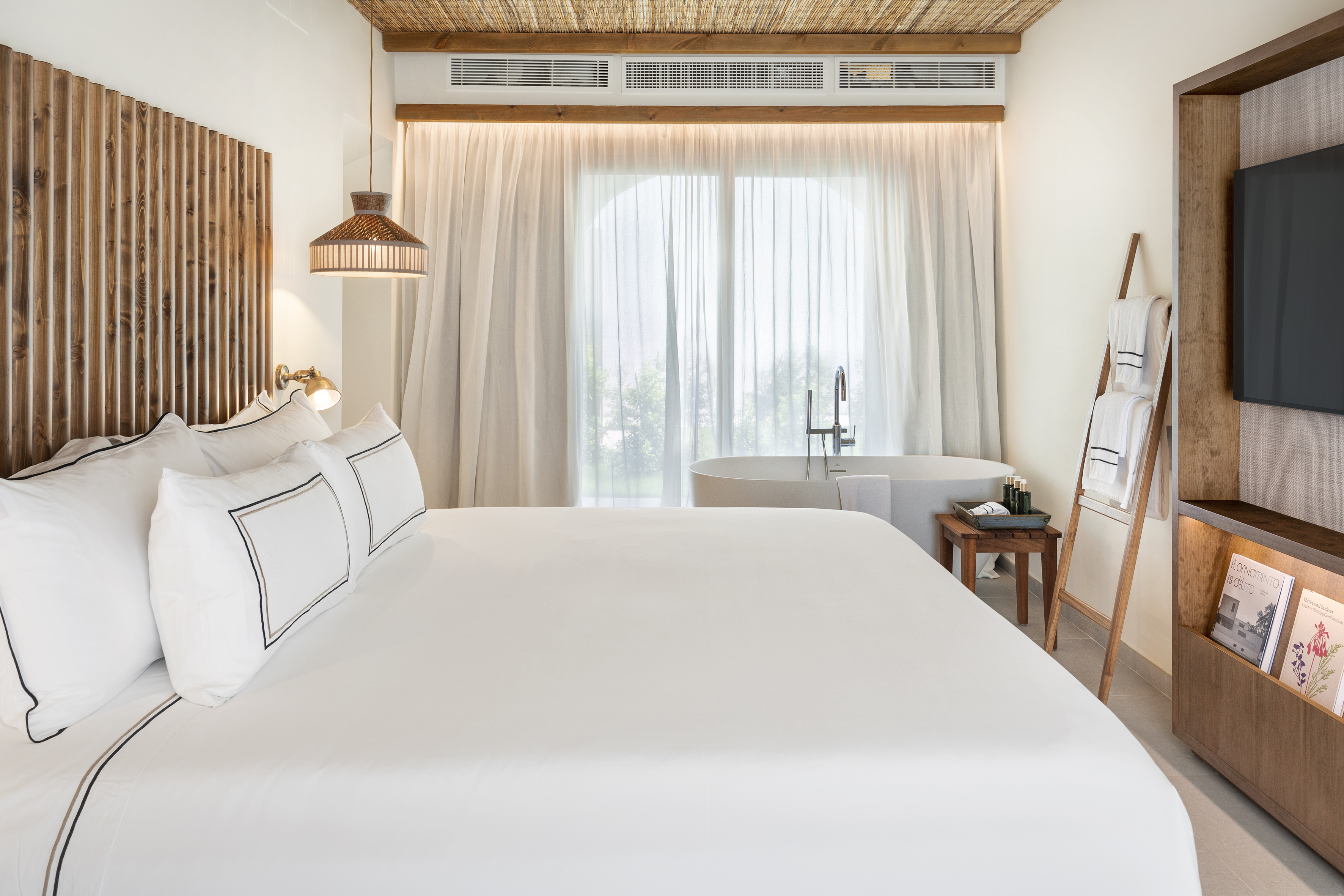 Modern hotel bedroom with white bed, wooden headboard, and freestanding bathtub.