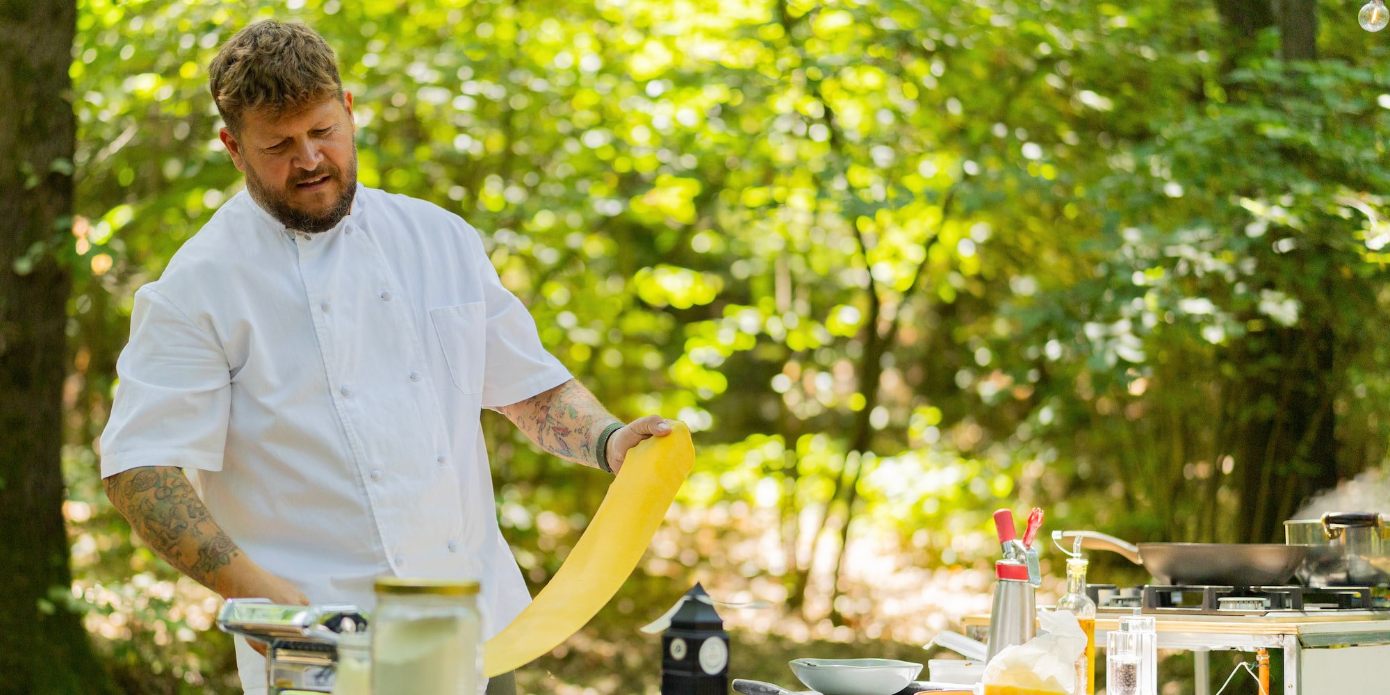 Chef making fresh pasta with a machine outdoors, surrounded by green trees.
