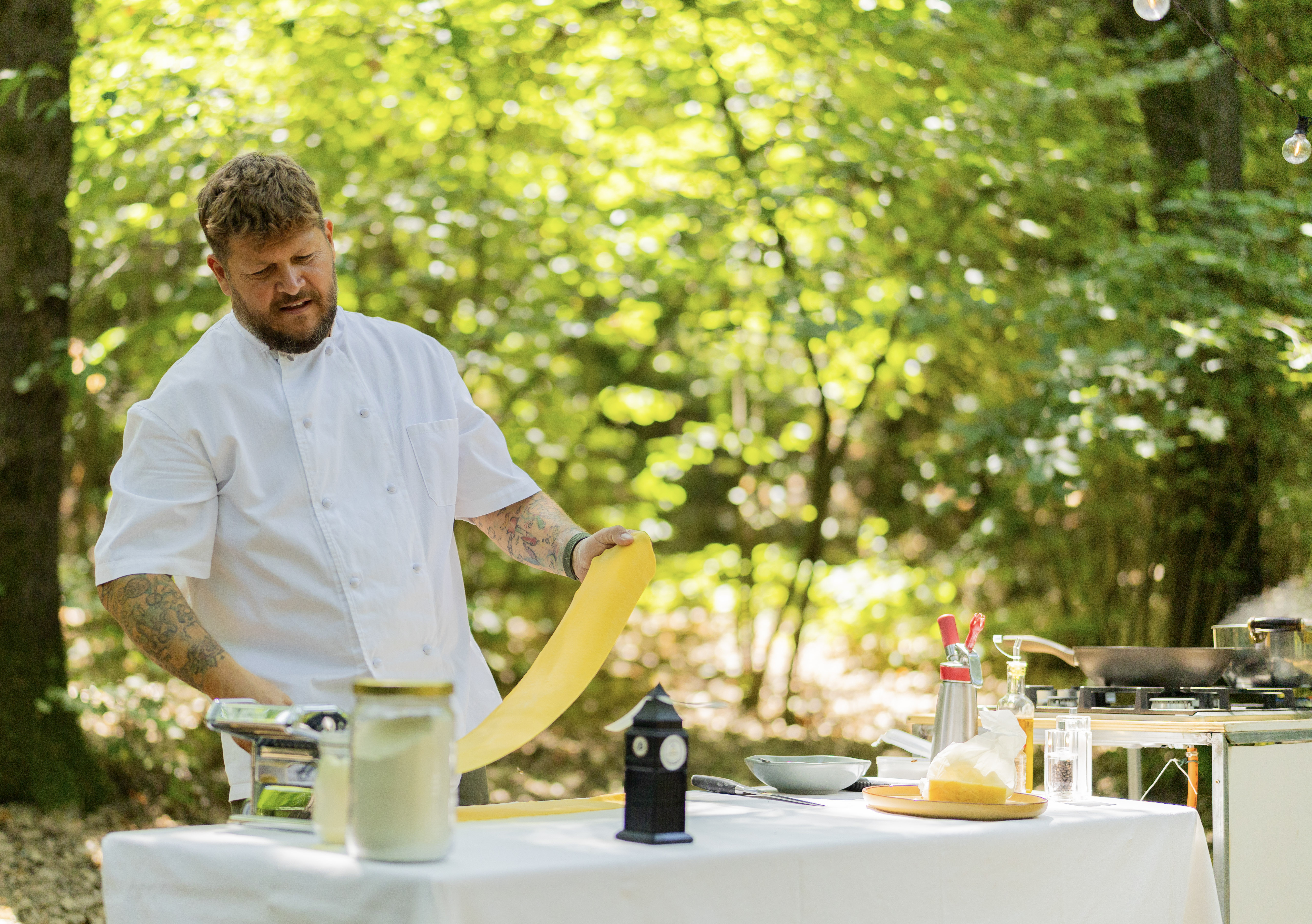 Chef making fresh pasta with a machine outdoors, surrounded by green trees.