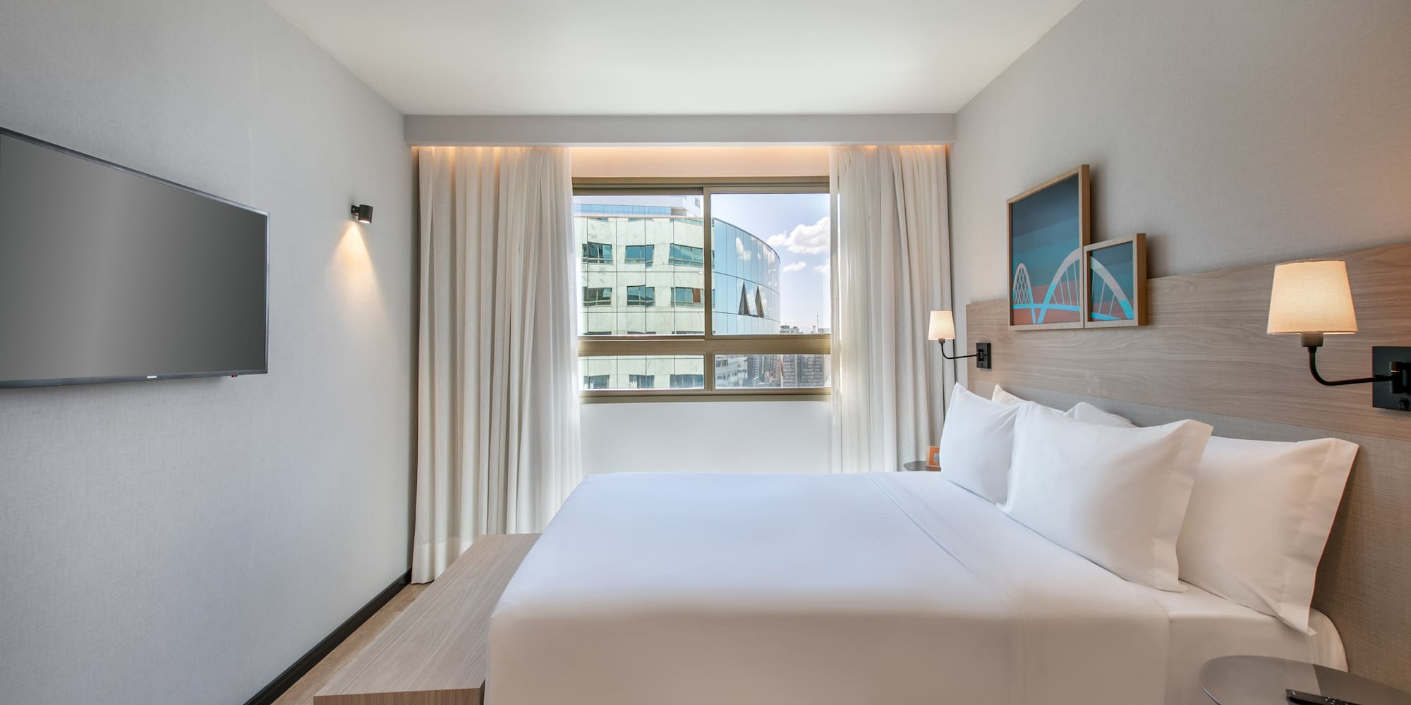 Modern hotel room with king bed, city view window, and wall-mounted TV.
