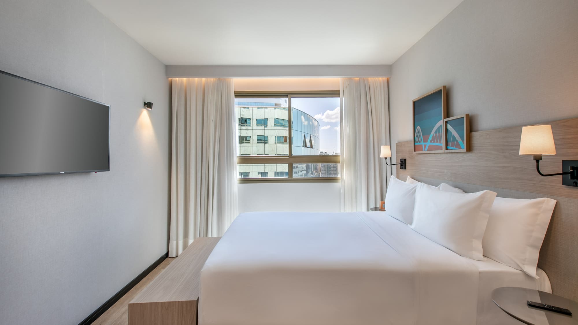 Modern hotel room with king bed, city view window, and wall-mounted TV.
