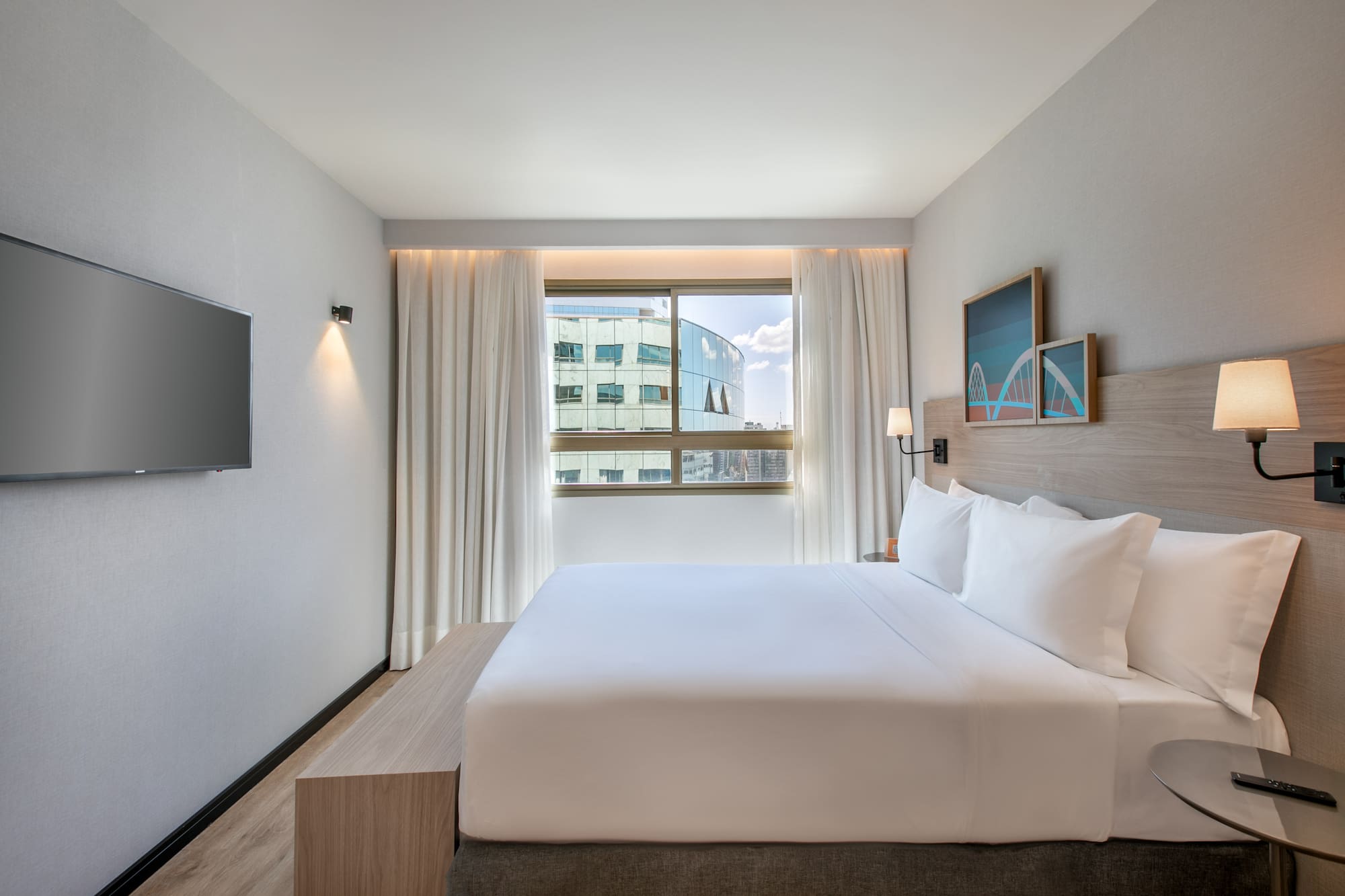 Modern hotel room with king bed, city view window, and wall-mounted TV.