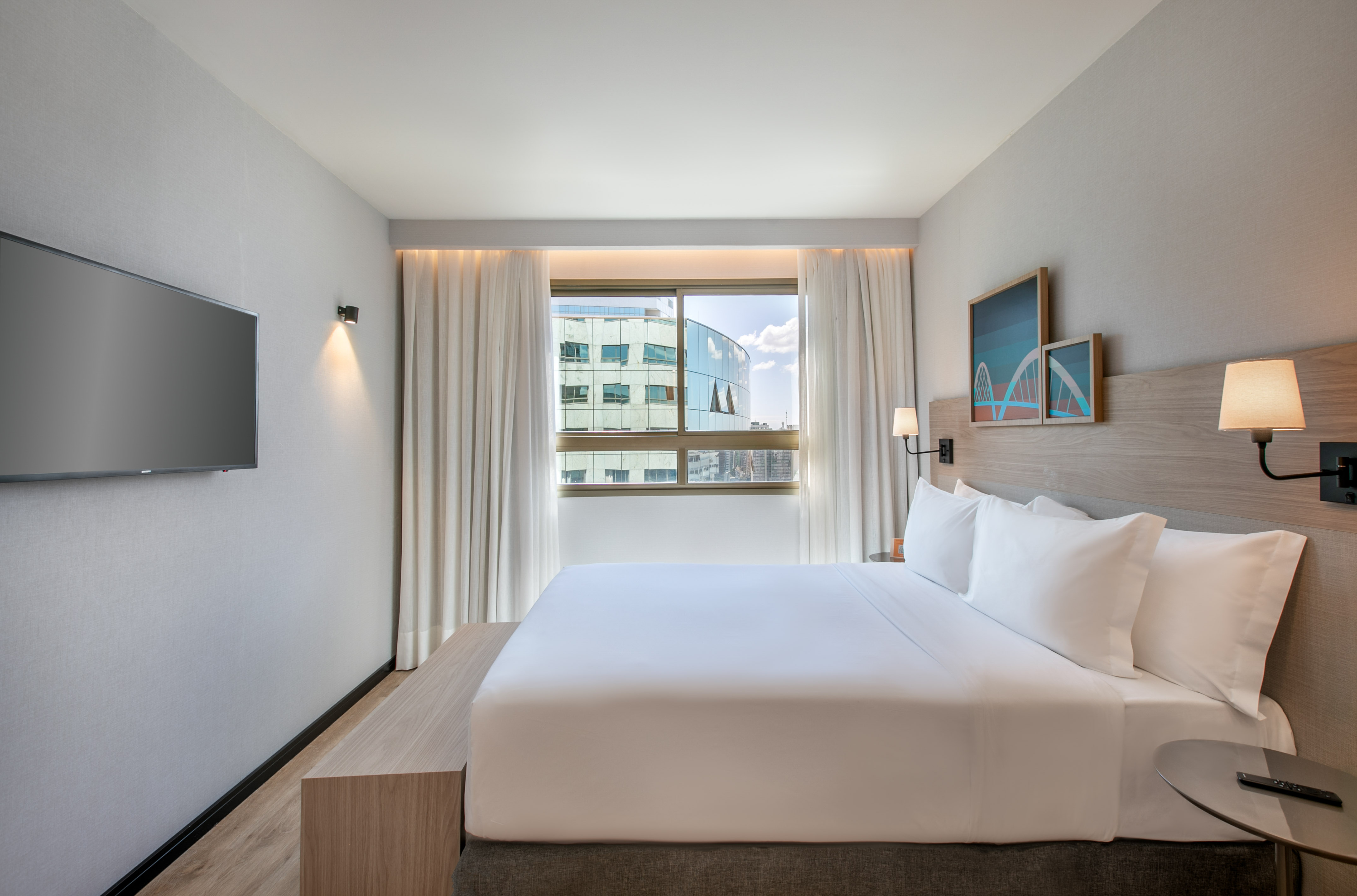 Modern hotel room with king bed, city view window, and wall-mounted TV.