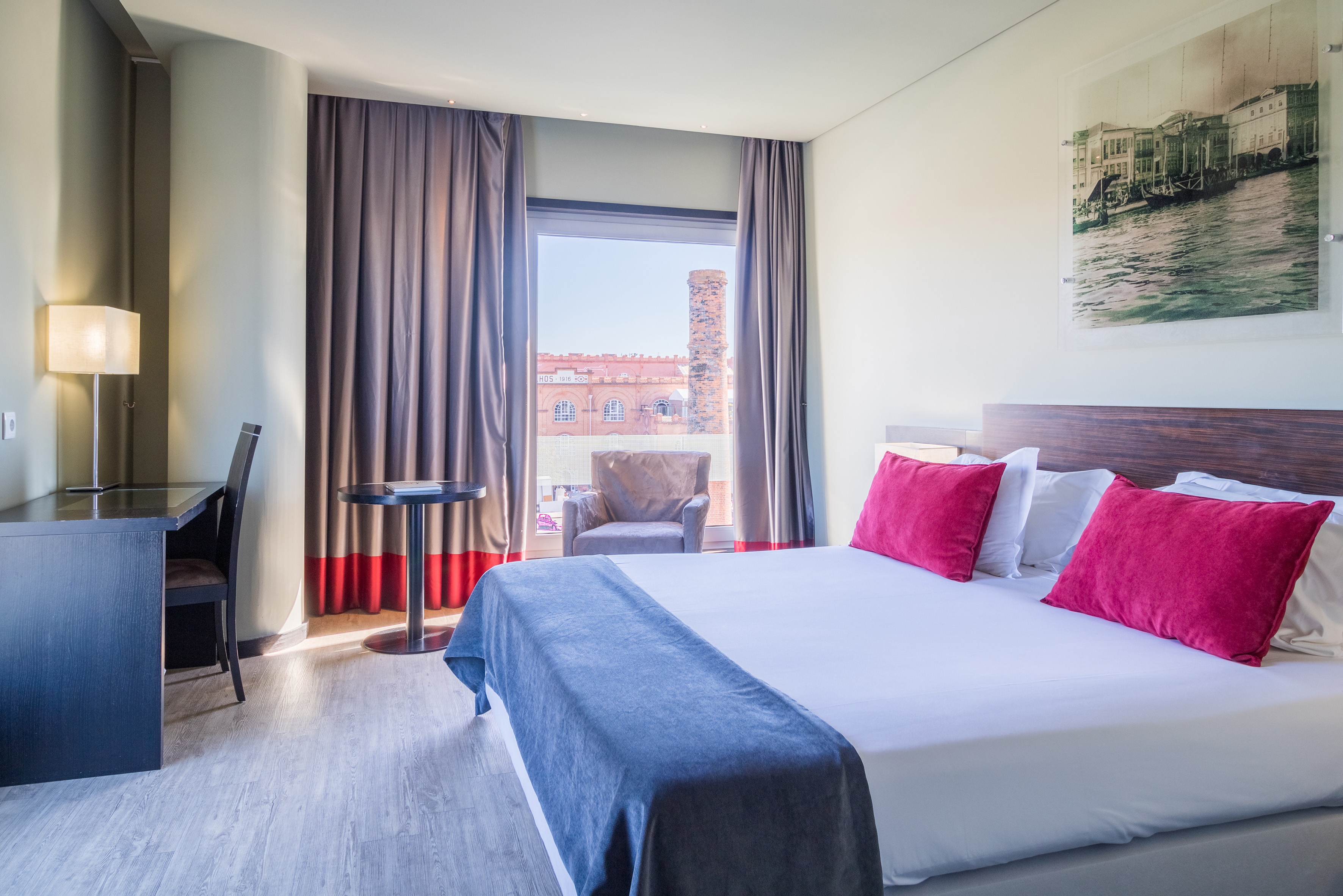 Modern hotel room with large bed, desk, city view, and magenta pillows.