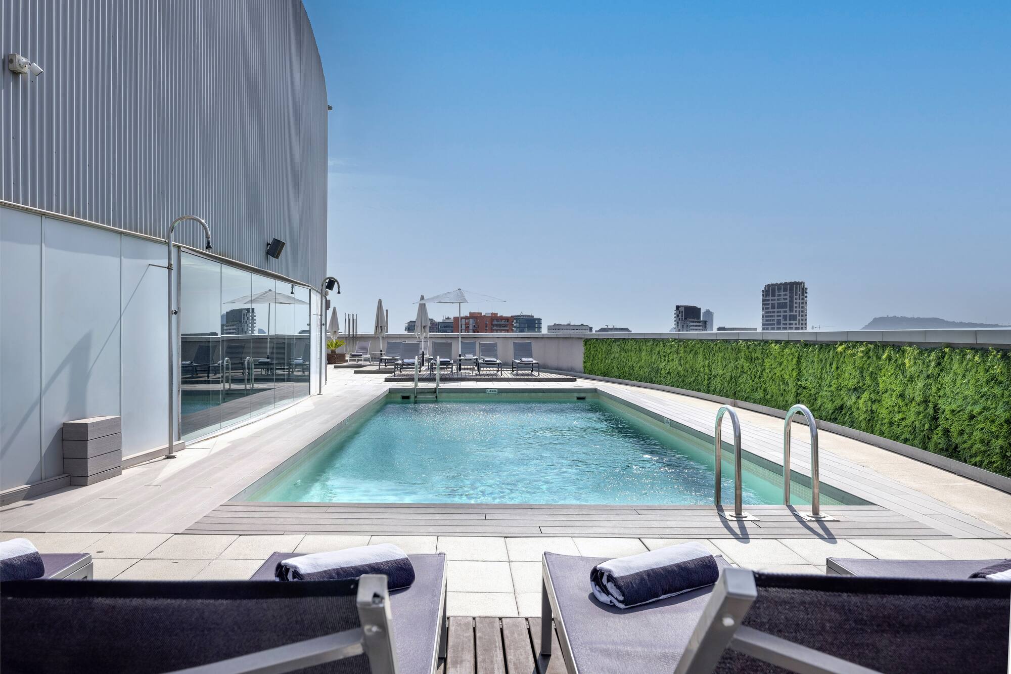 Rooftop hotel swimming pool with lounge chairs, city view, and blue sky.
