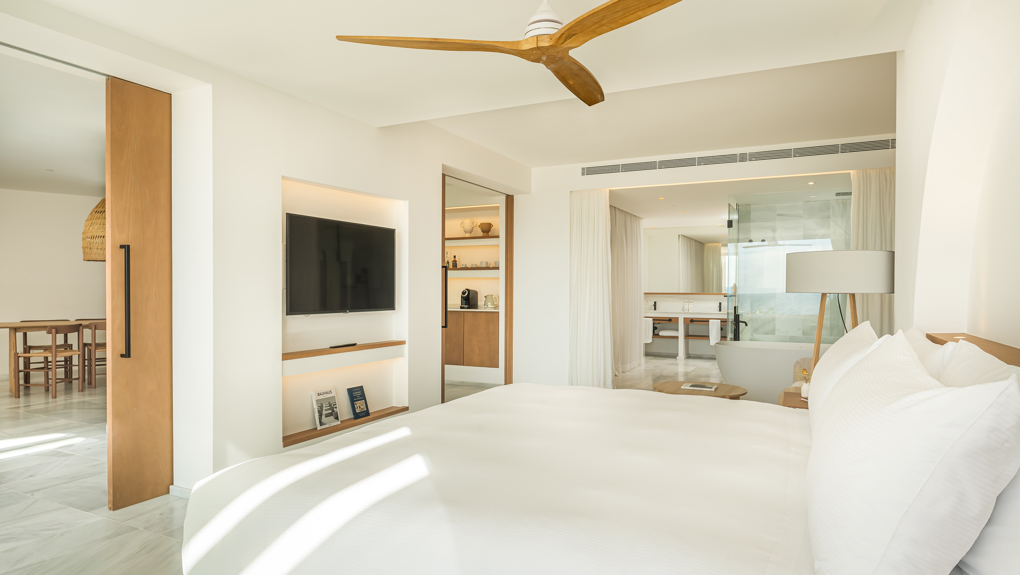 Luxurious modern hotel suite with white bed, wooden fan, and open bathroom.