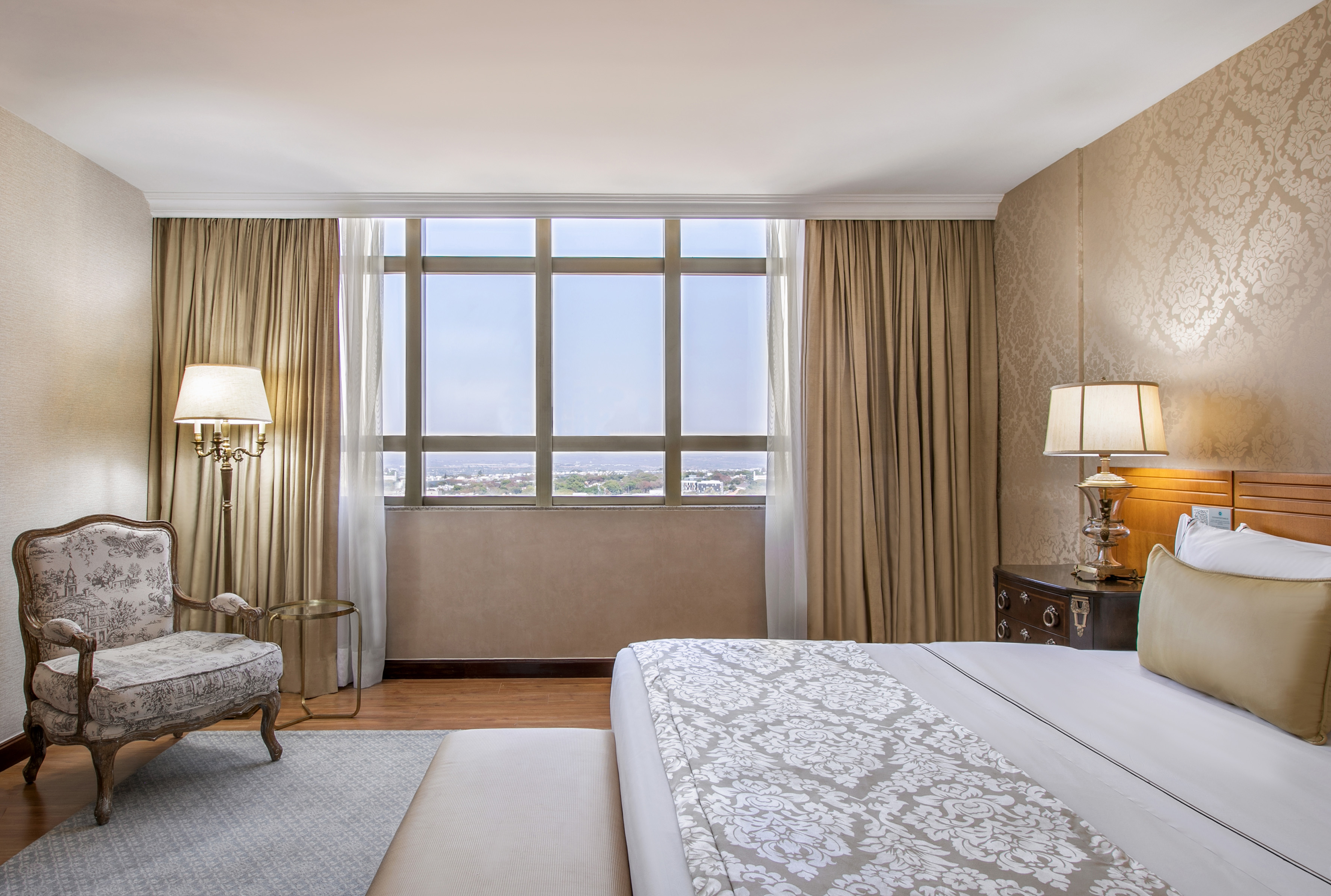 Hotel king bedroom with elegant armchair, large window, and city view.