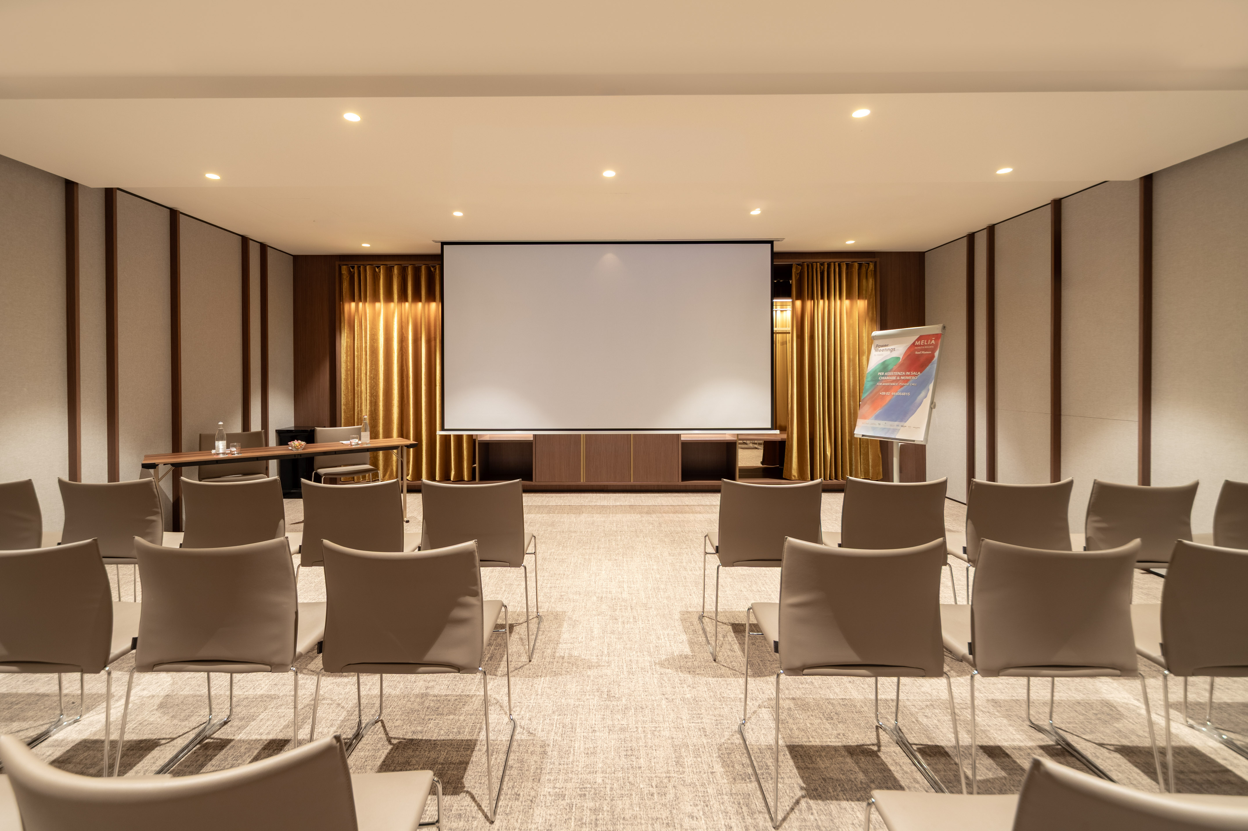 Hotel meeting room with projection screen, empty chairs, and warm presentation setup.