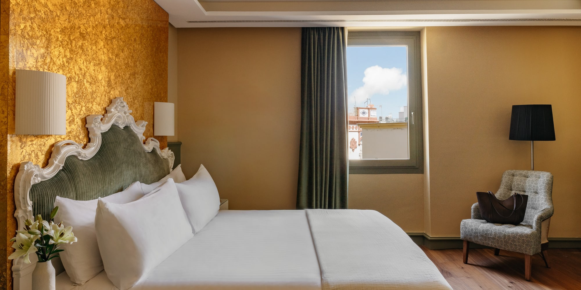 Dormitorio elegante de hotel con cama, cabecero clásico, pared dorada y ventana.