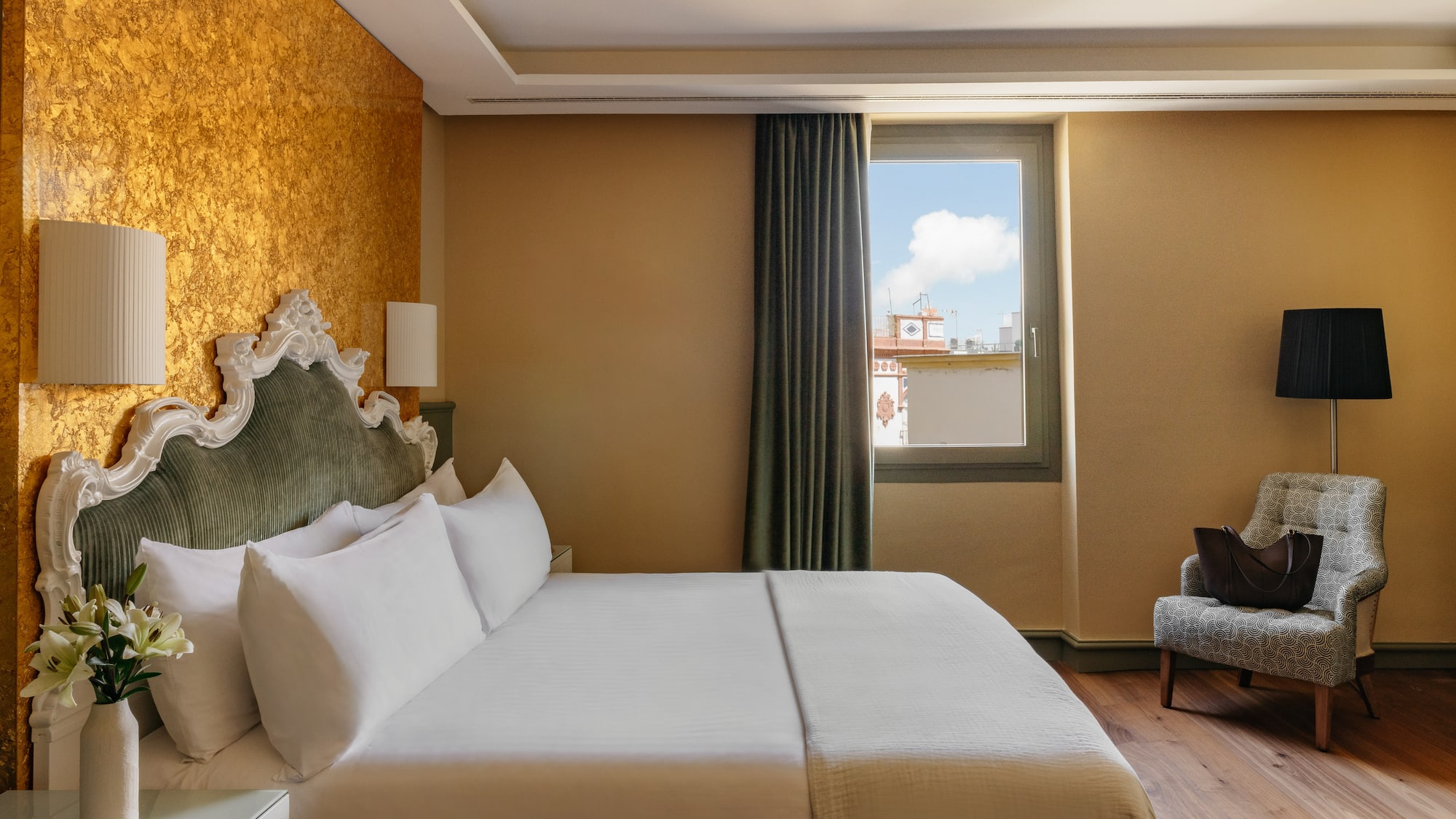 Dormitorio elegante de hotel con cama, cabecero clásico, pared dorada y ventana.