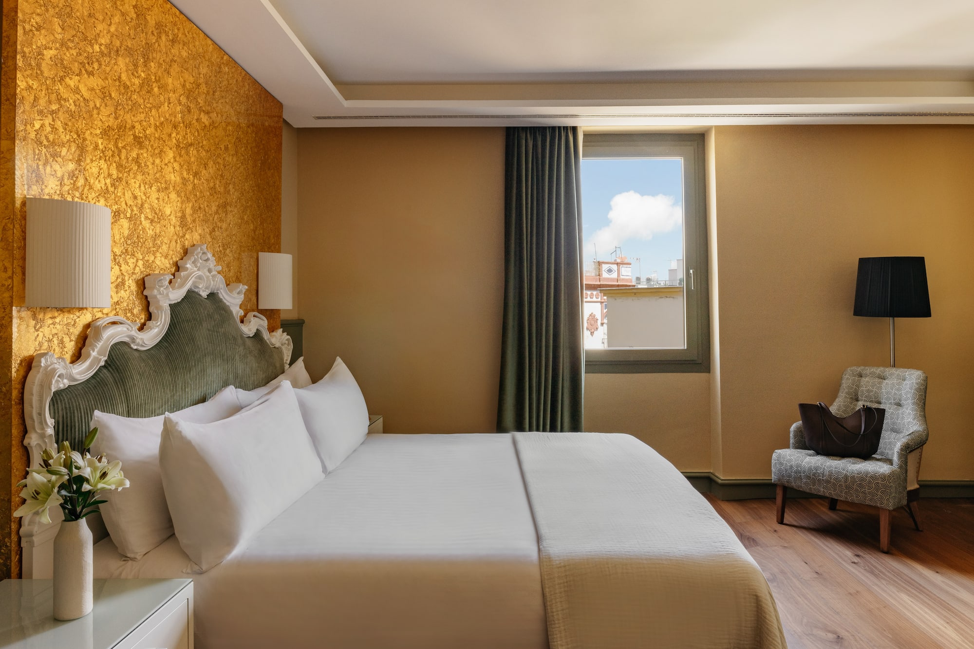 Dormitorio elegante de hotel con cama, cabecero clásico, pared dorada y ventana.