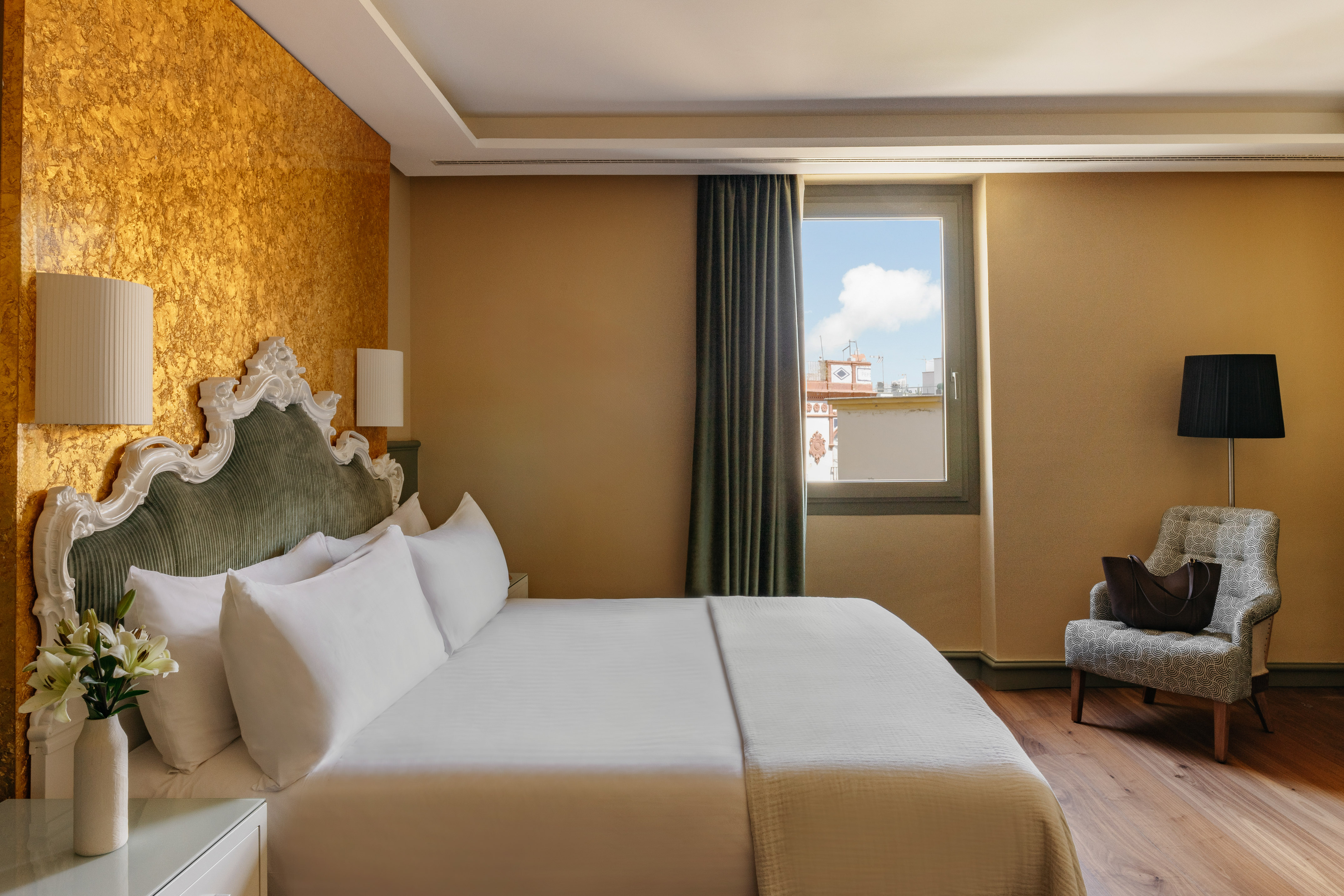 Dormitorio elegante de hotel con cama, cabecero clásico, pared dorada y ventana.