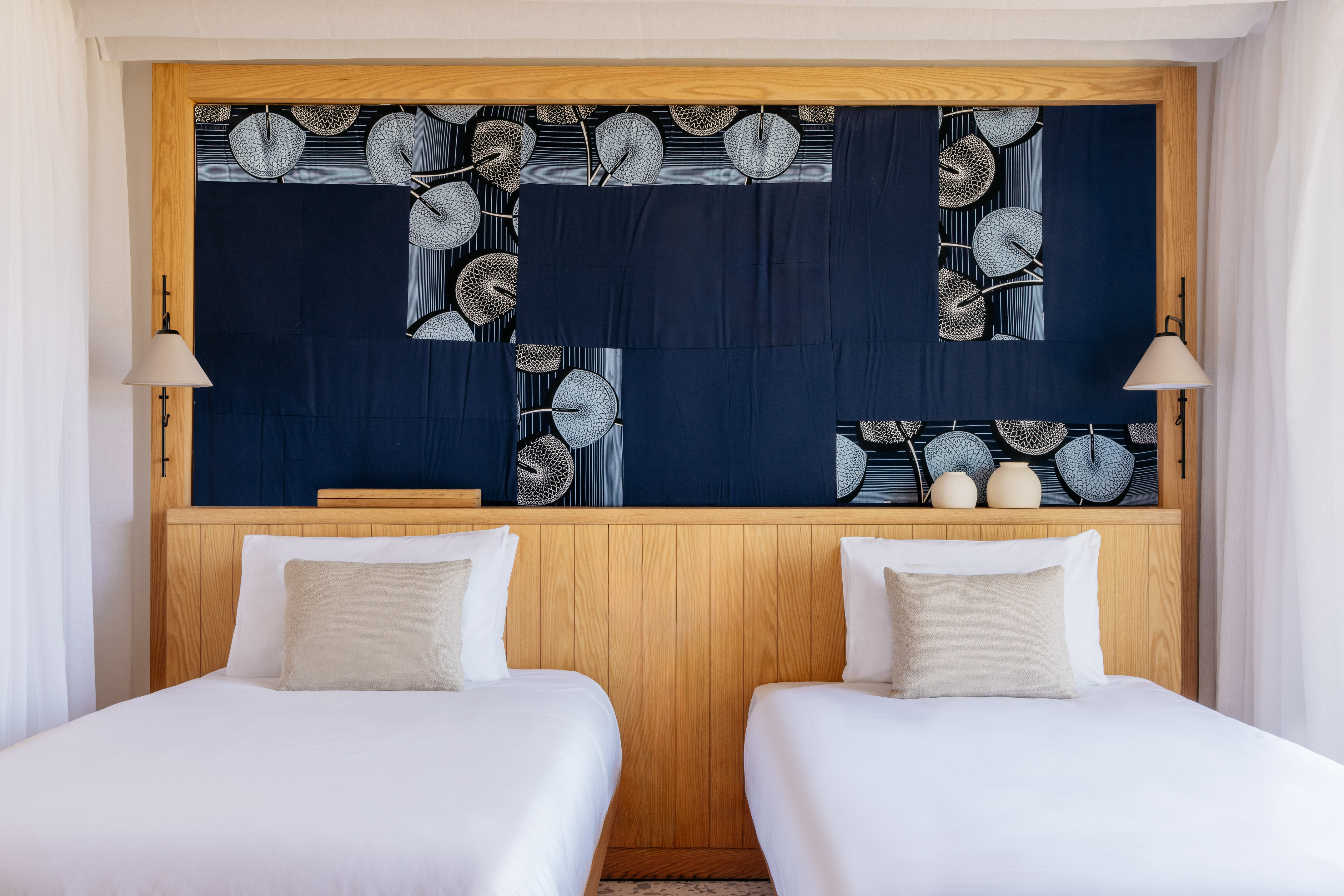 Modern hotel room: twin beds, wooden headboard, blue patterned fabric art.