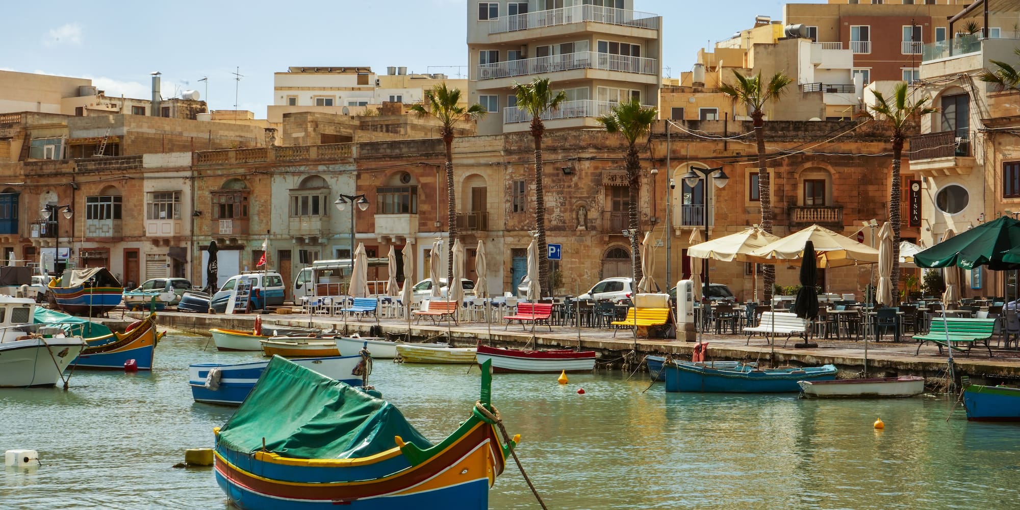 Malta harbor with colorful boats, waterfront buildings, cafes, and palm trees.