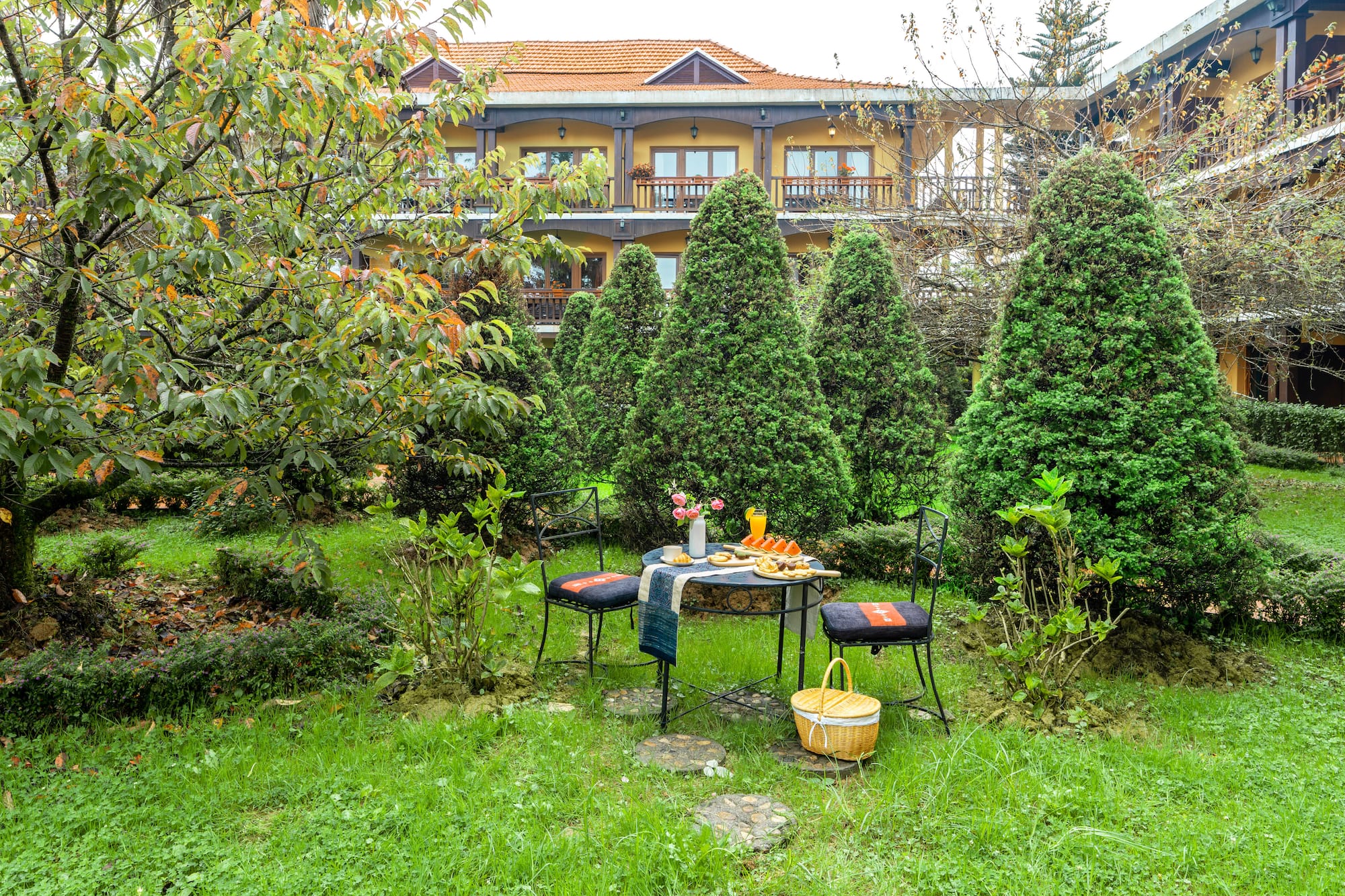 Garden patio breakfast table with lush evergreens, yellow hotel building background.