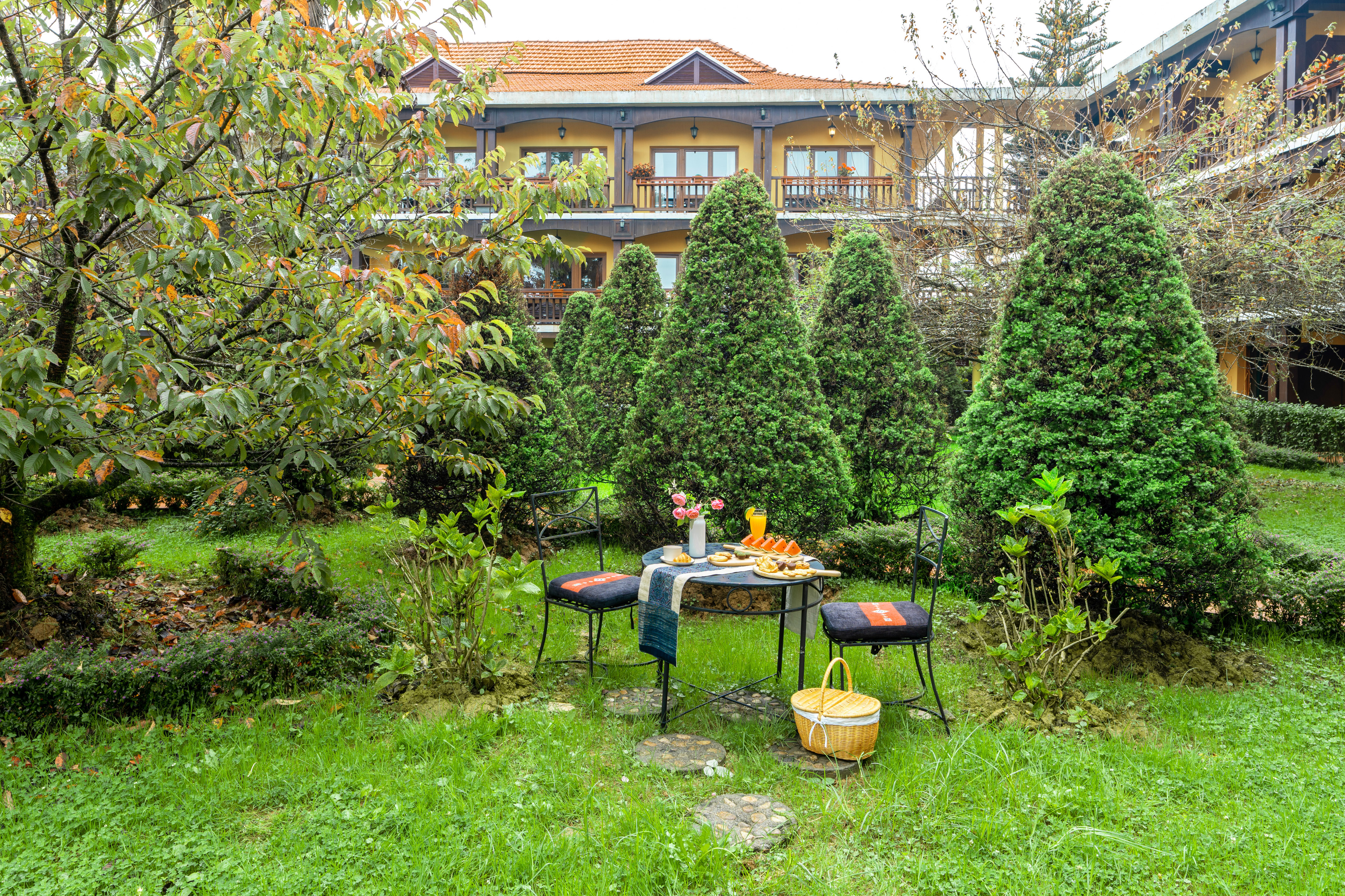 Garden patio breakfast table with lush evergreens, yellow hotel building background.
