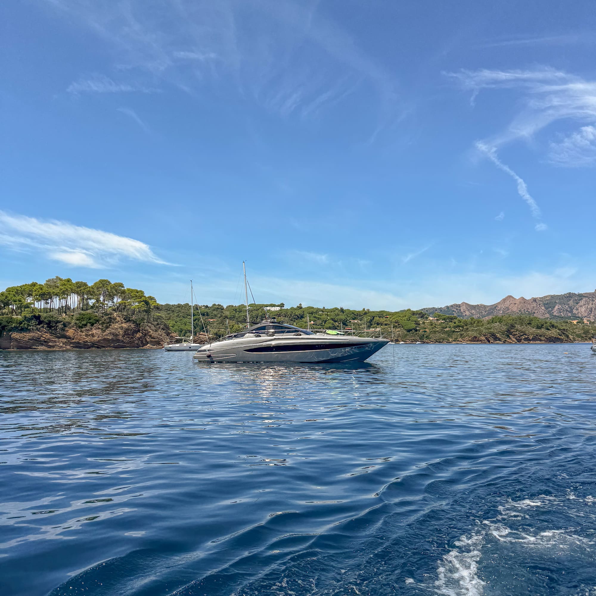 Luxury grey yacht on sparkling blue sea with green wooded coast and clear sky.