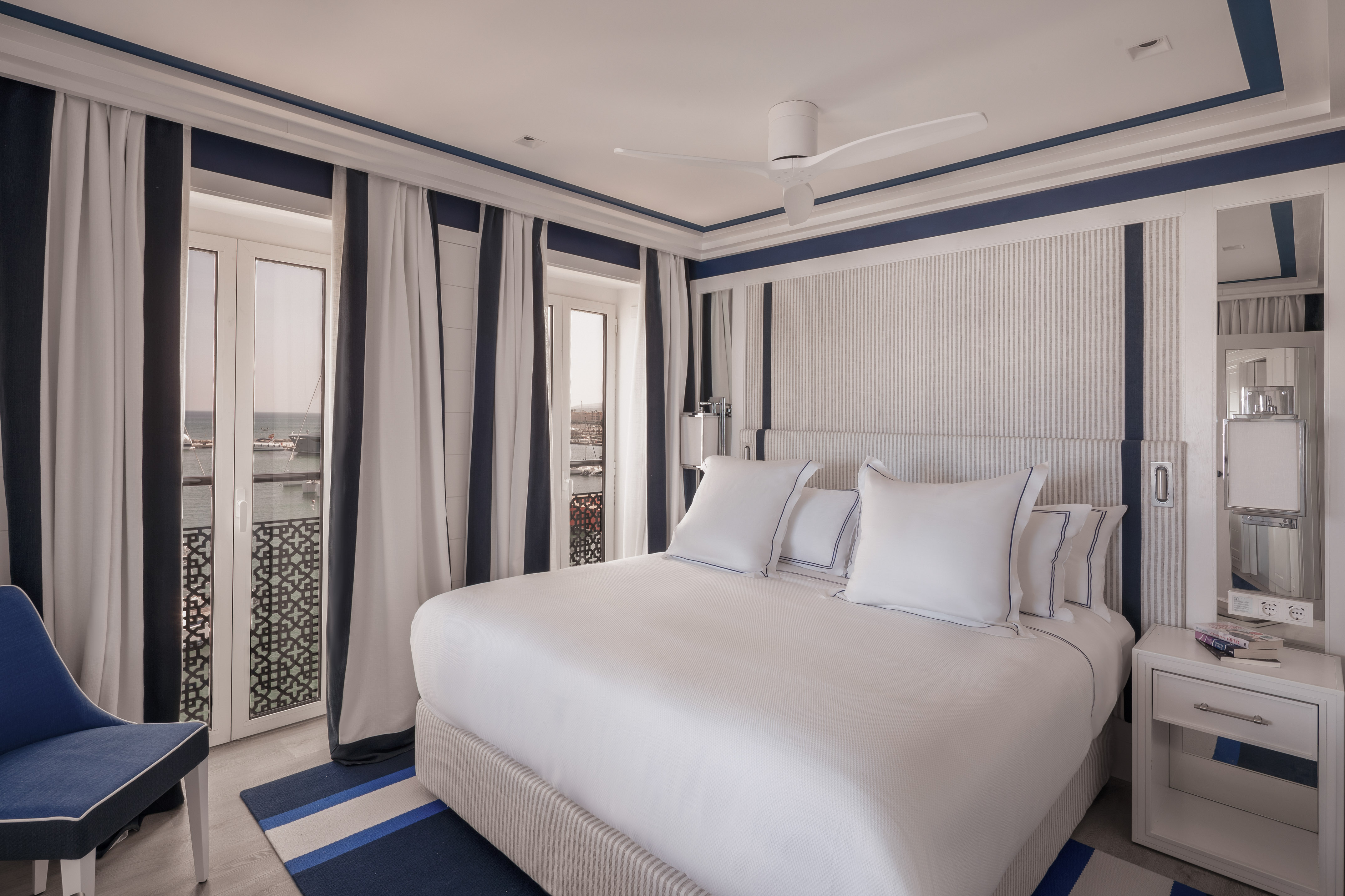 Luxury hotel bedroom with white and blue nautical decor, large bed, and sea view balcony.