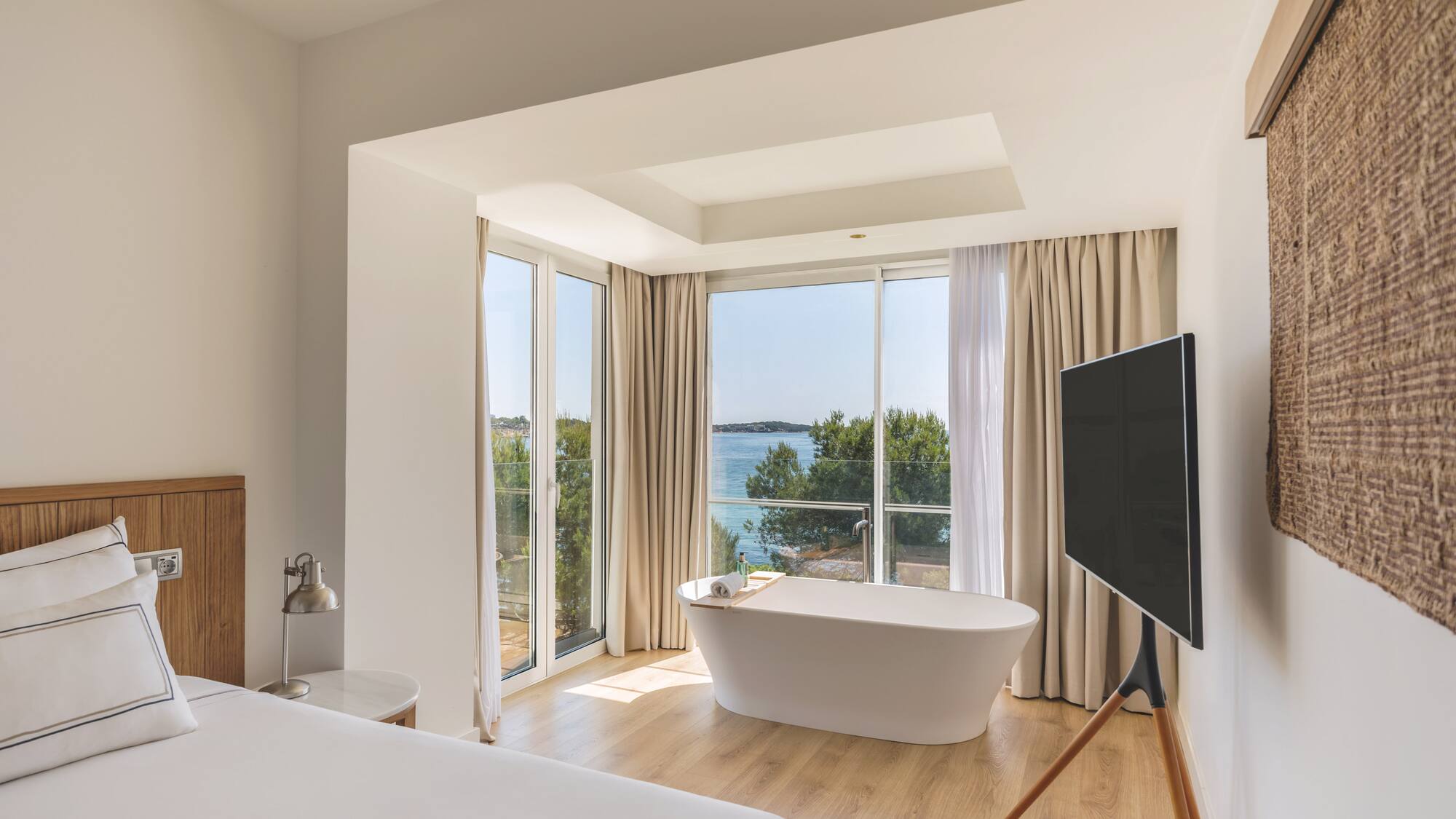 Modern hotel room with sea view, freestanding bathtub, and white bed.