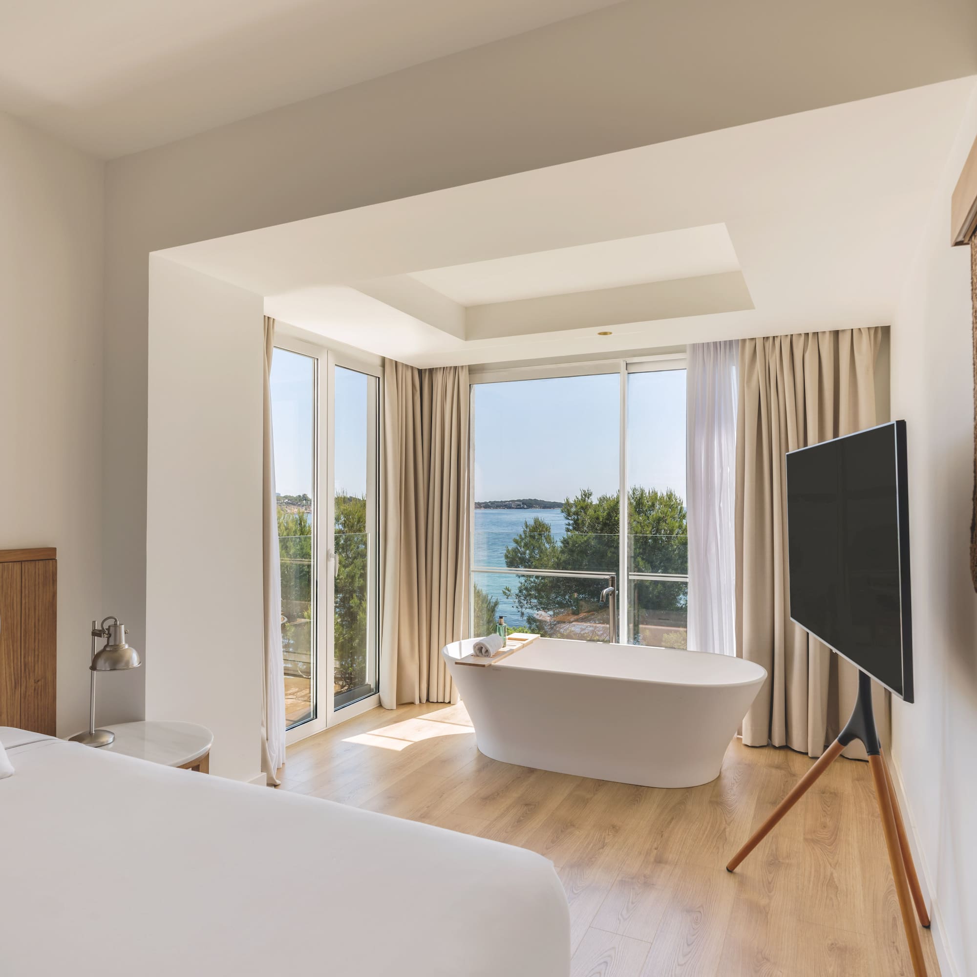 Modern hotel room with sea view, freestanding bathtub, and white bed.