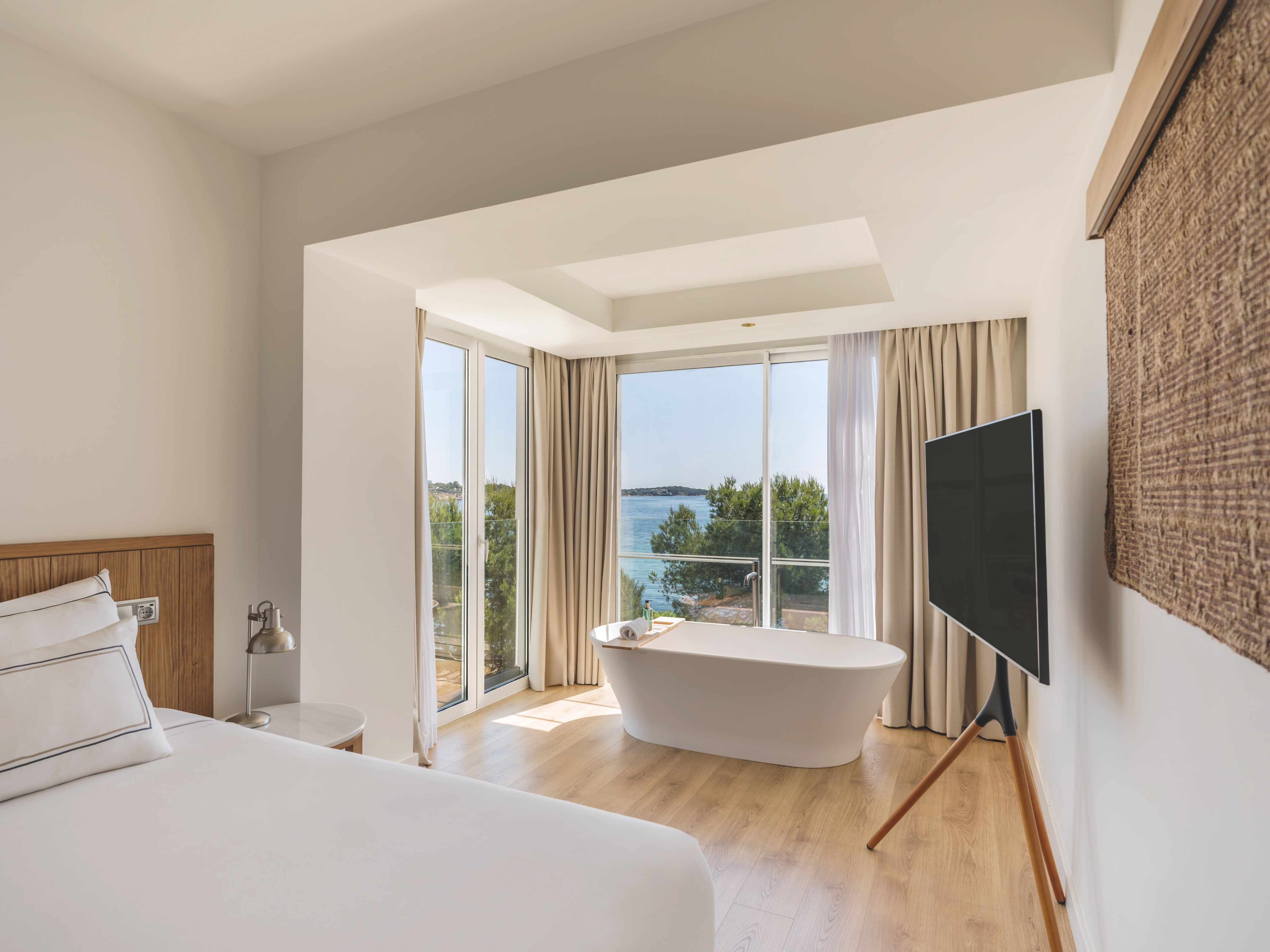 Modern hotel room with sea view, freestanding bathtub, and white bed.