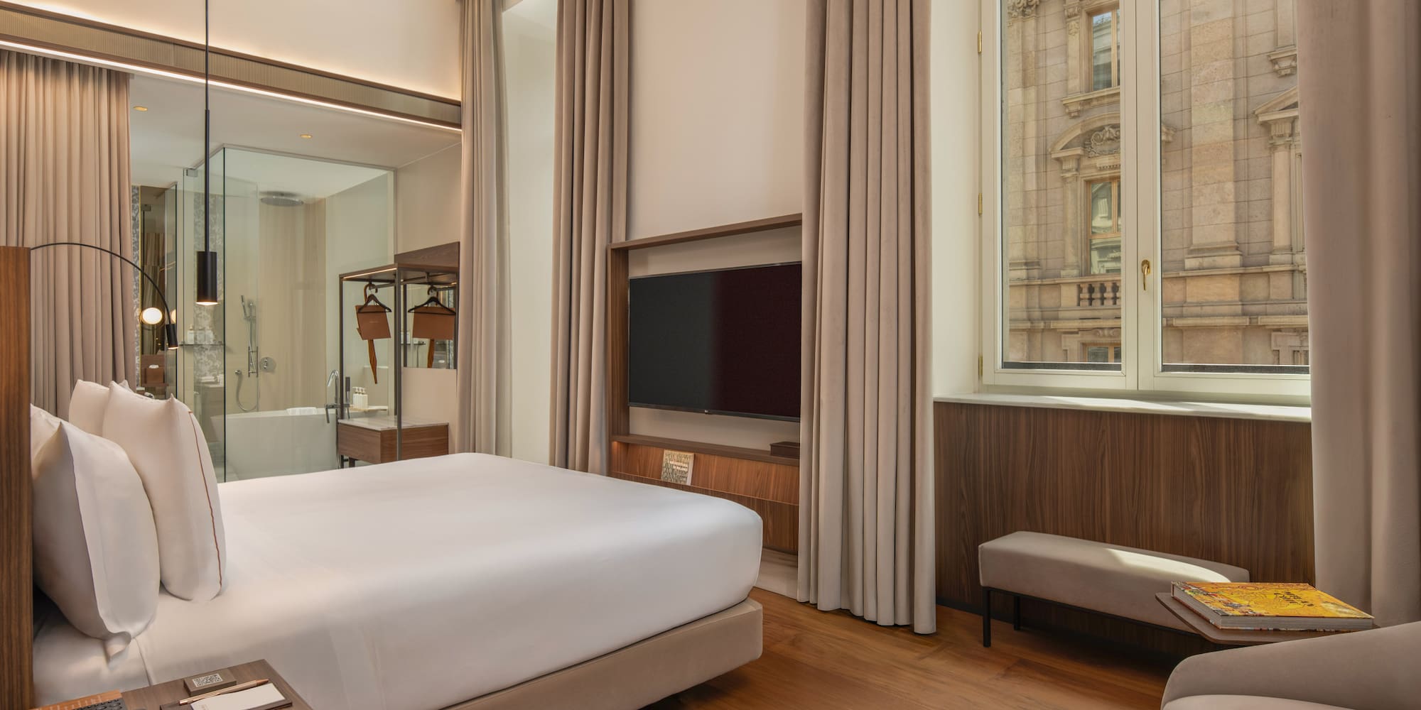 Modern hotel room with king bed, wood floor, city view, glass bathroom.