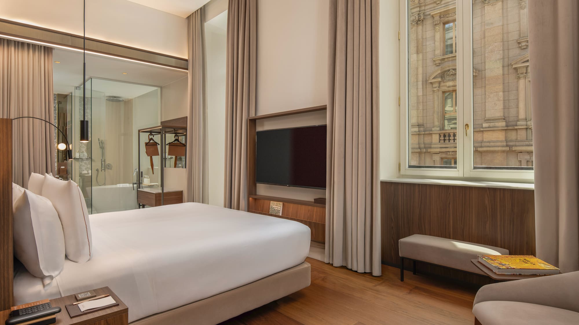 Modern hotel room with king bed, wood floor, city view, glass bathroom.