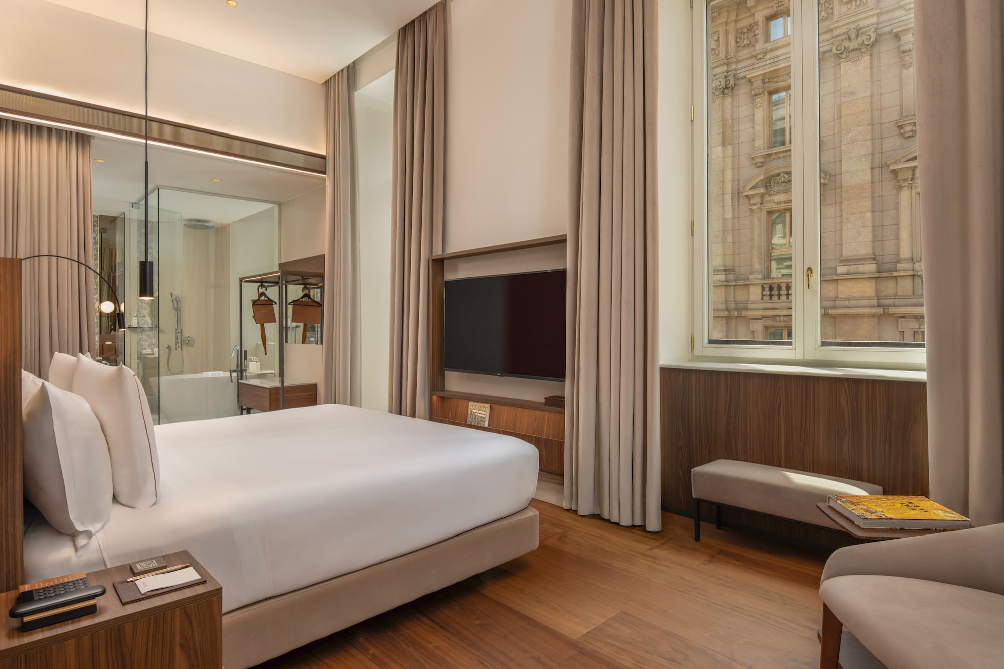 Modern hotel room with king bed, wood floor, city view, glass bathroom.