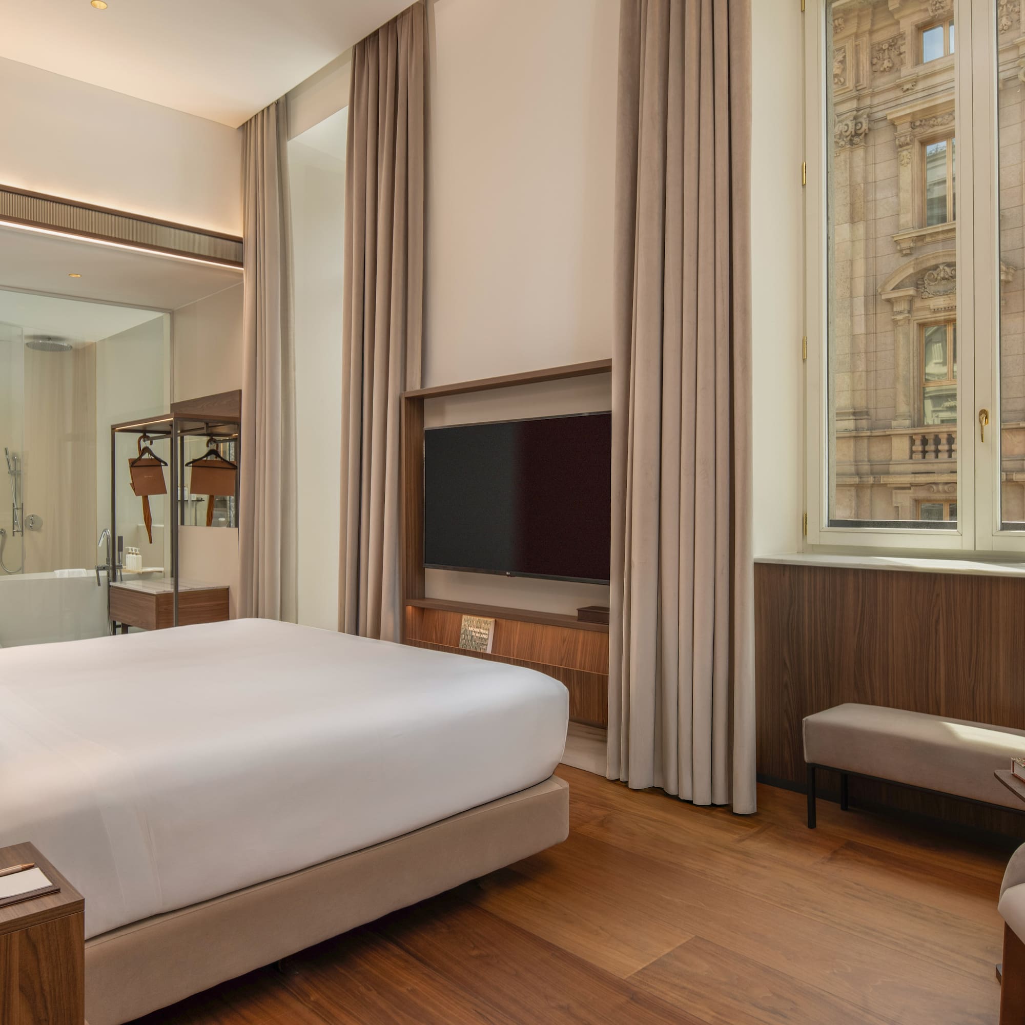 Modern hotel room with king bed, wood floor, city view, glass bathroom.