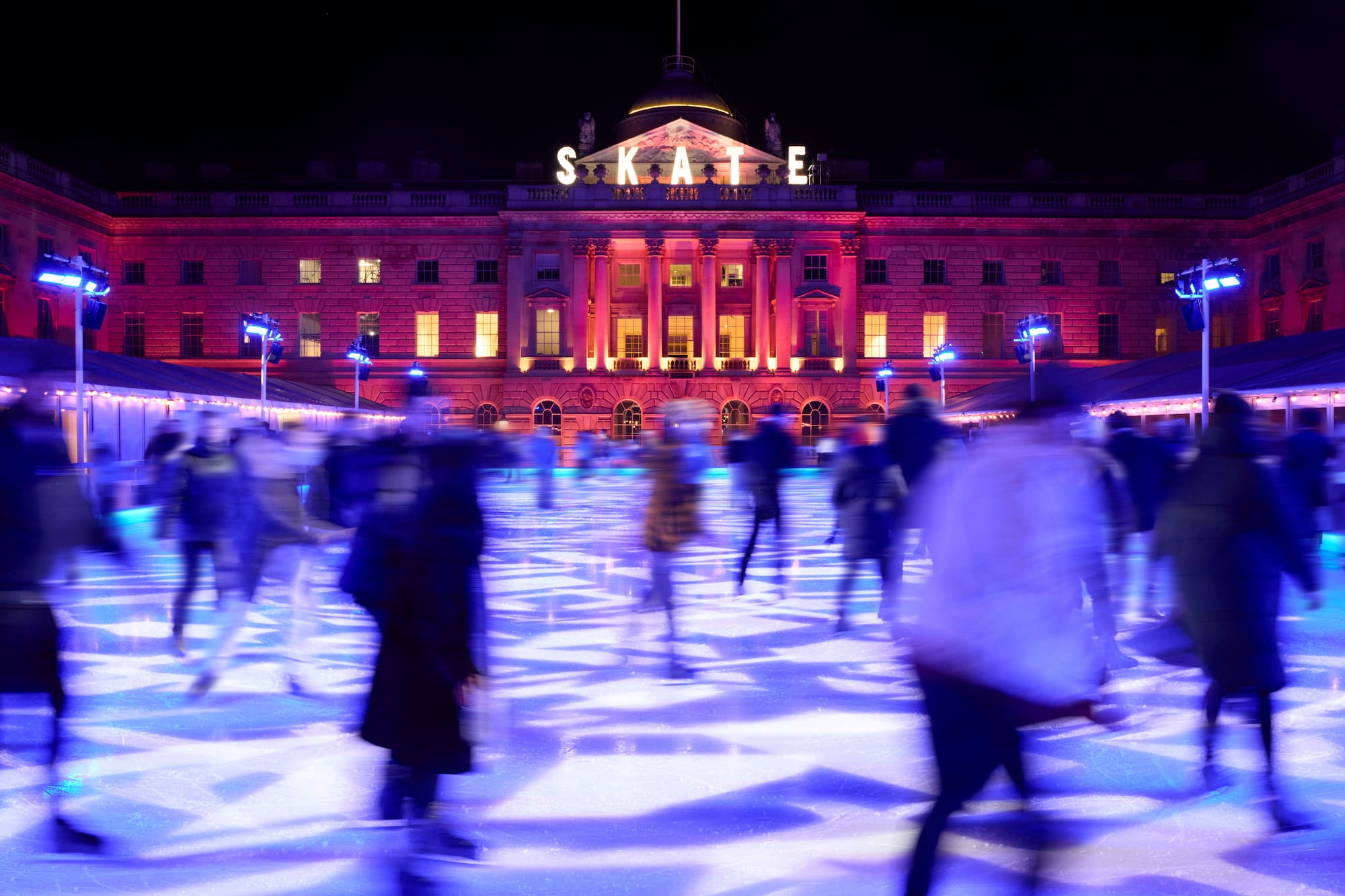 Pista de hielo SKATE nocturna con patinadores, luces azules y edificio rosa.