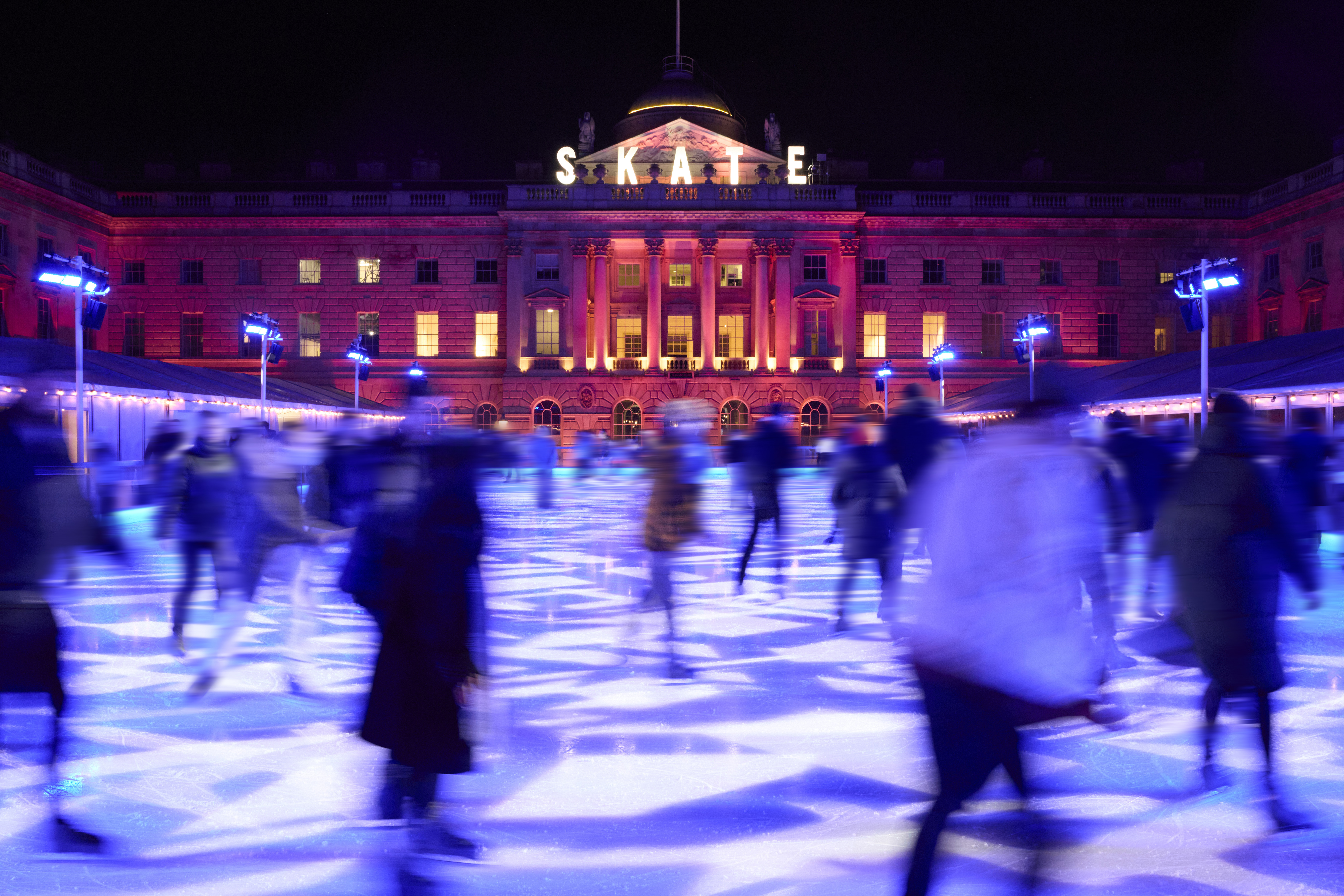Pista de hielo SKATE nocturna con patinadores, luces azules y edificio rosa.