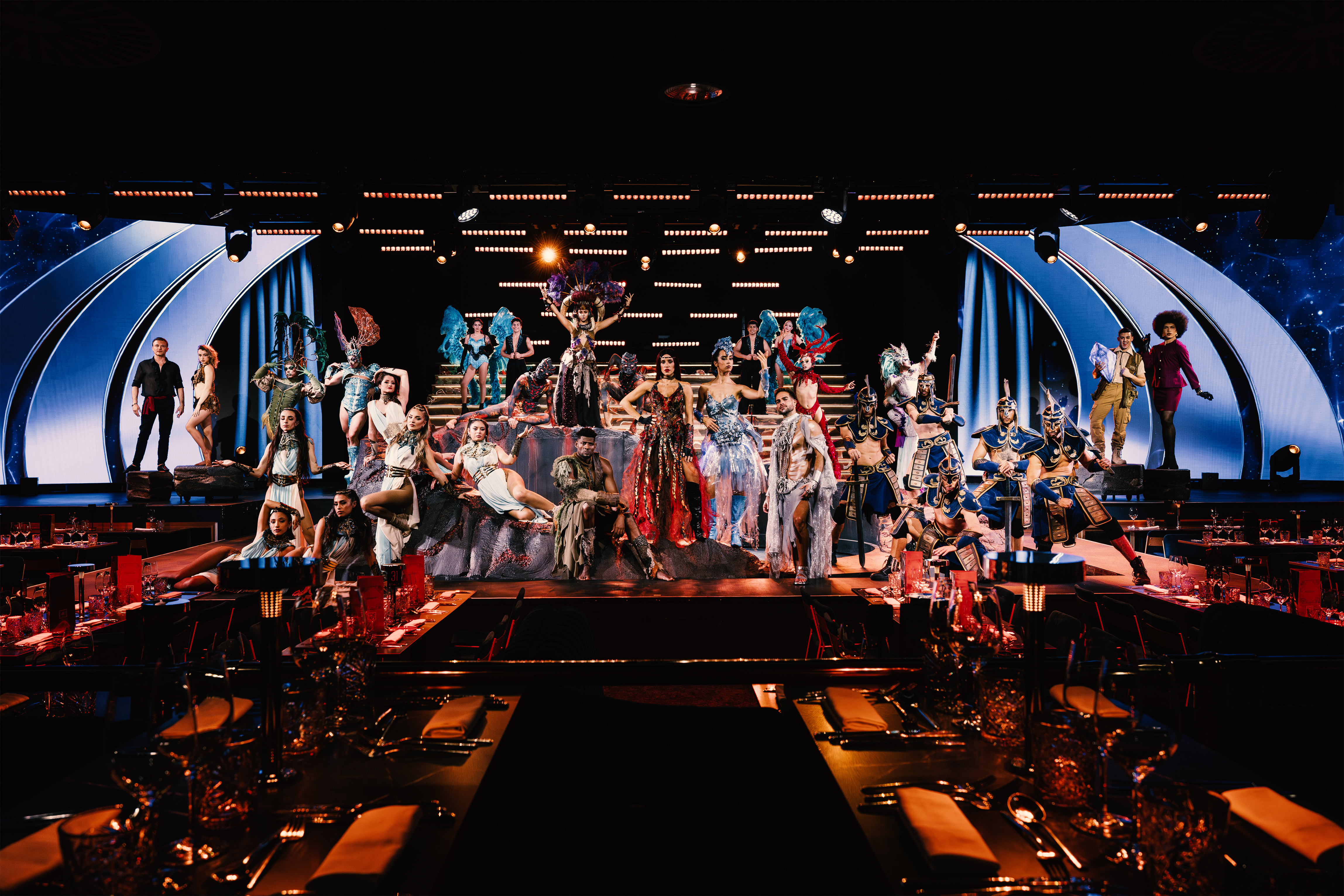 Diverse performers in vibrant costumes on a dramatic stage for a dinner show.