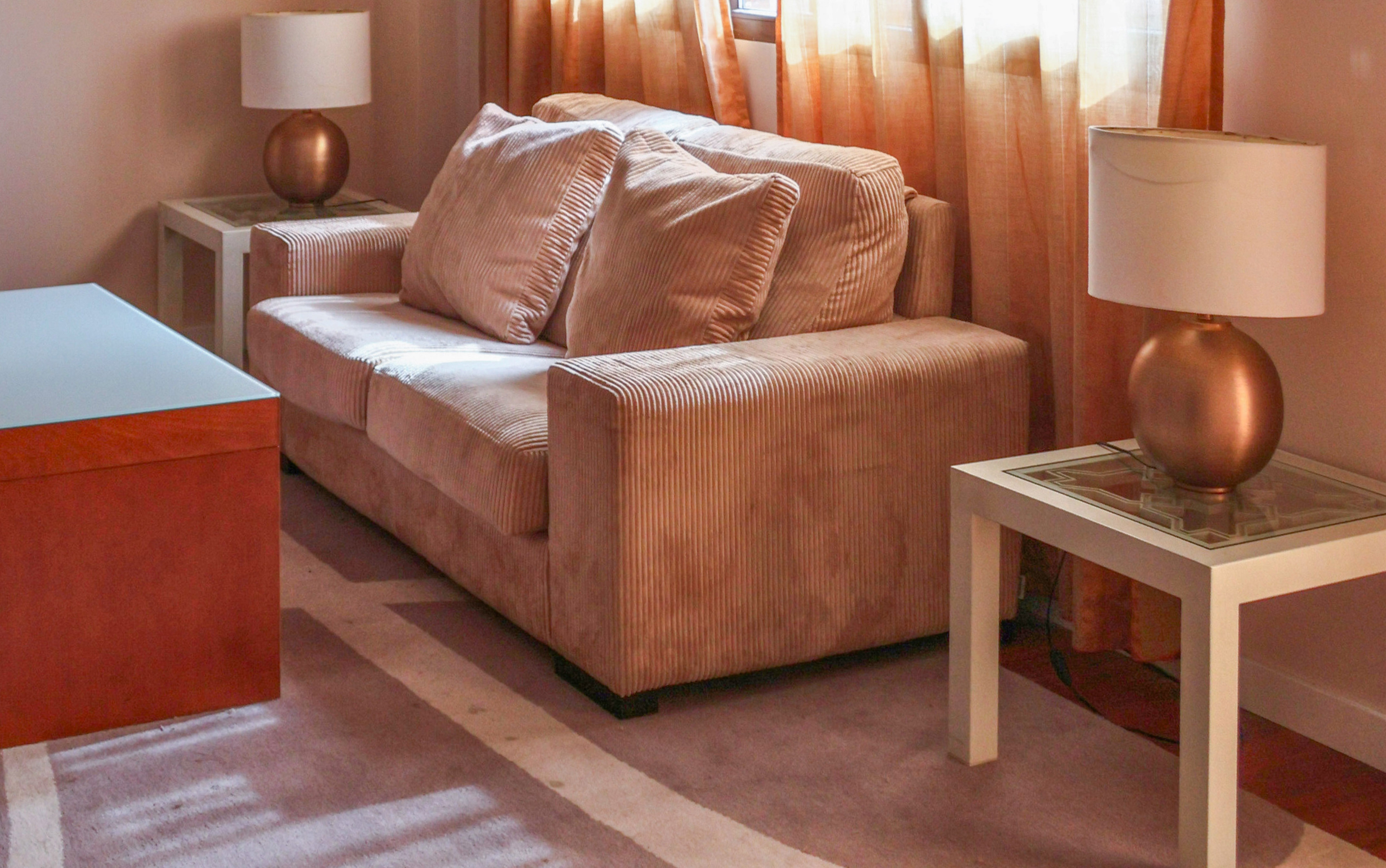Beige corduroy sofa in a cozy living room with lamps, coffee table, and orange curtains.