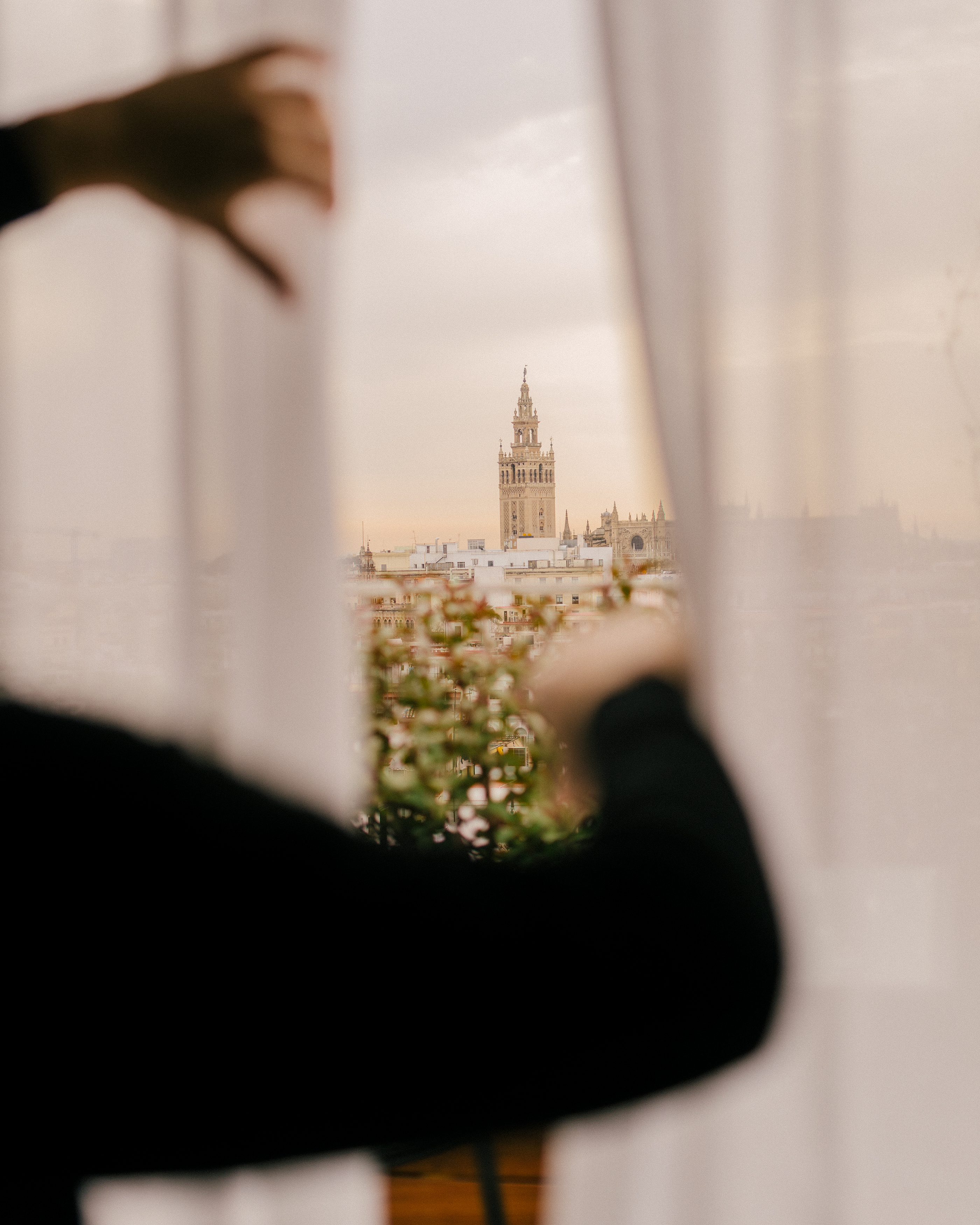 Hands opening curtains revealing Giralda tower and Seville city view.