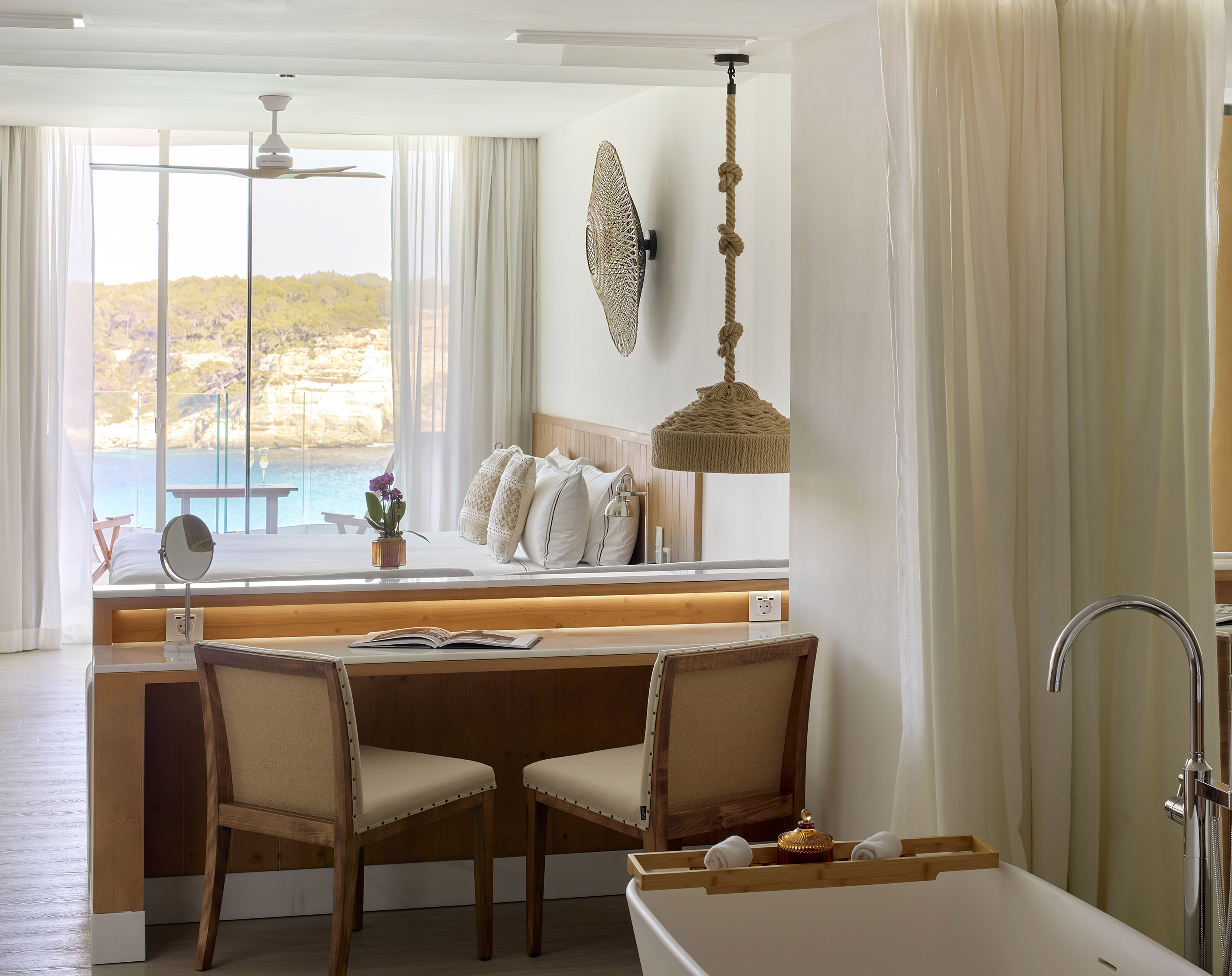 Bright hotel suite with panoramic sea view, bed, desk, and luxury tub.