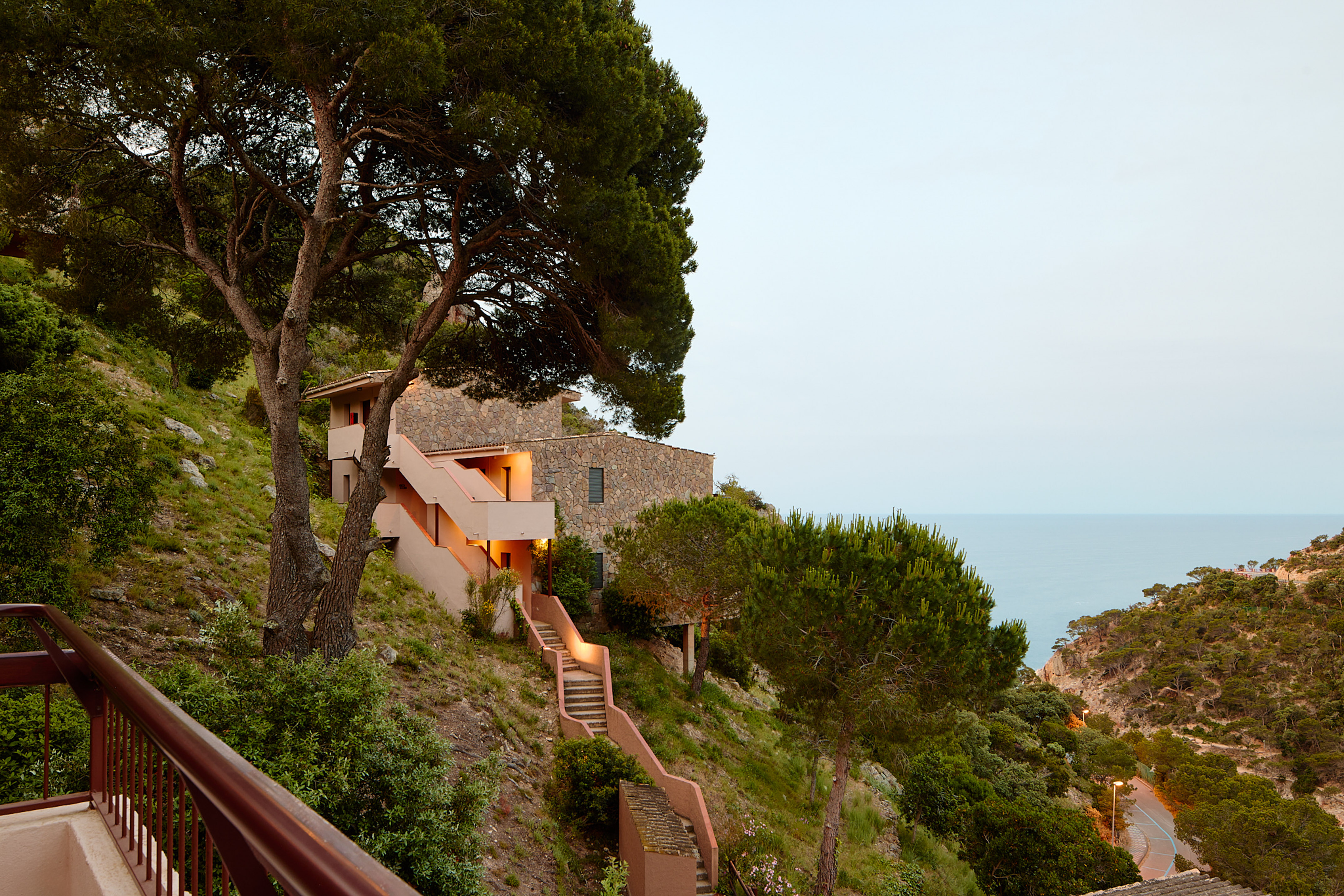 Coastal stone house on green hillside with terraced steps and ocean view.