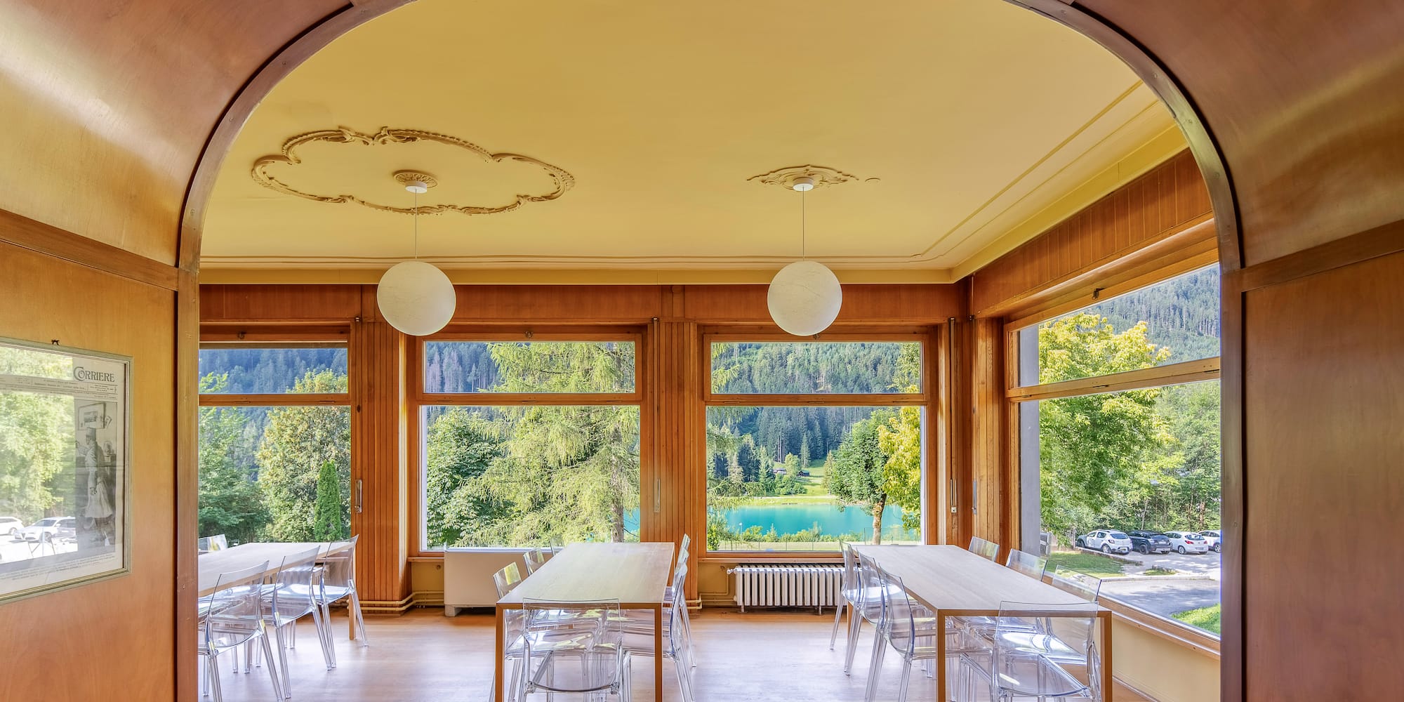 Dining room overlooking a blue lake, forest, and mountains with clear chairs.