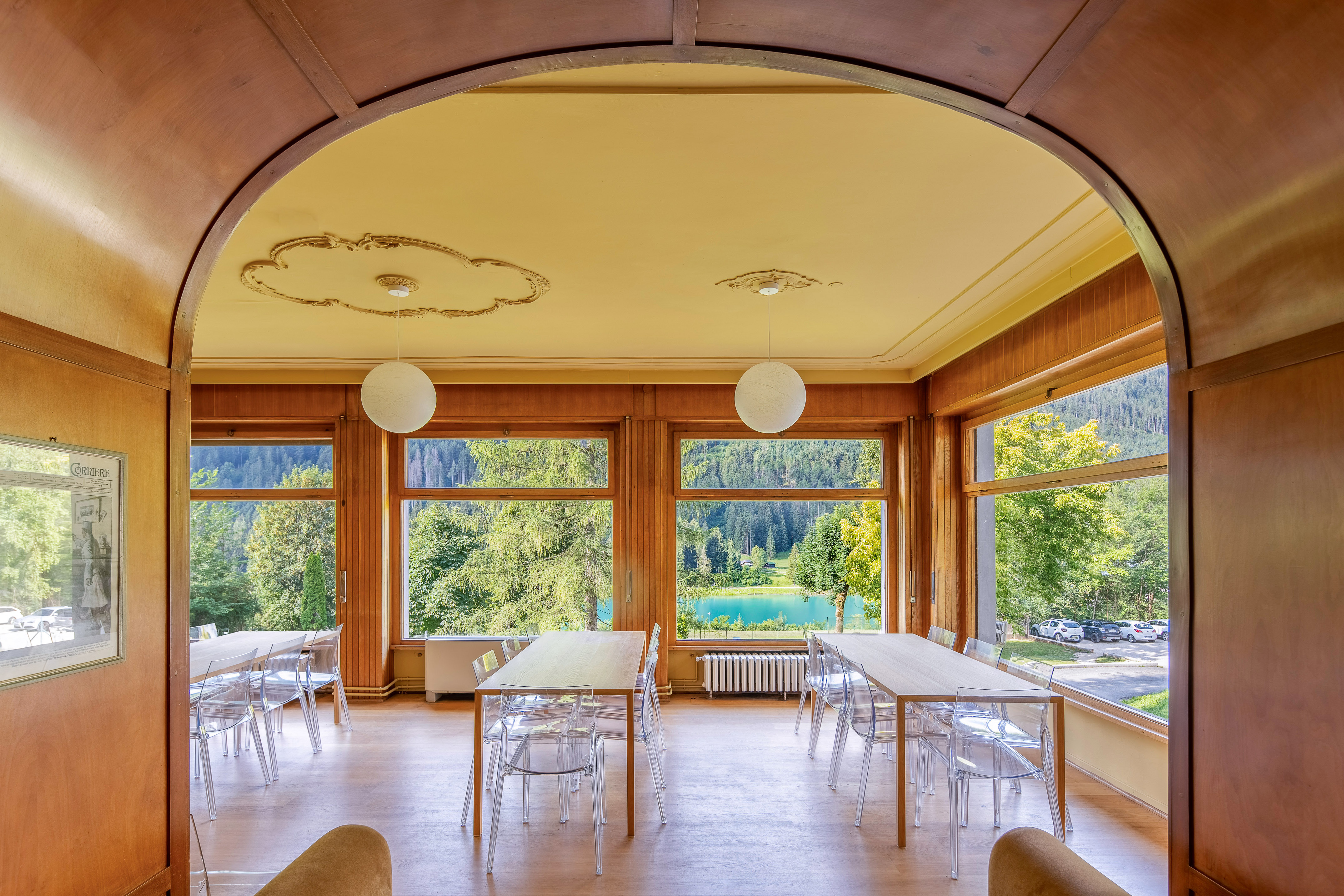 Dining room overlooking a blue lake, forest, and mountains with clear chairs.