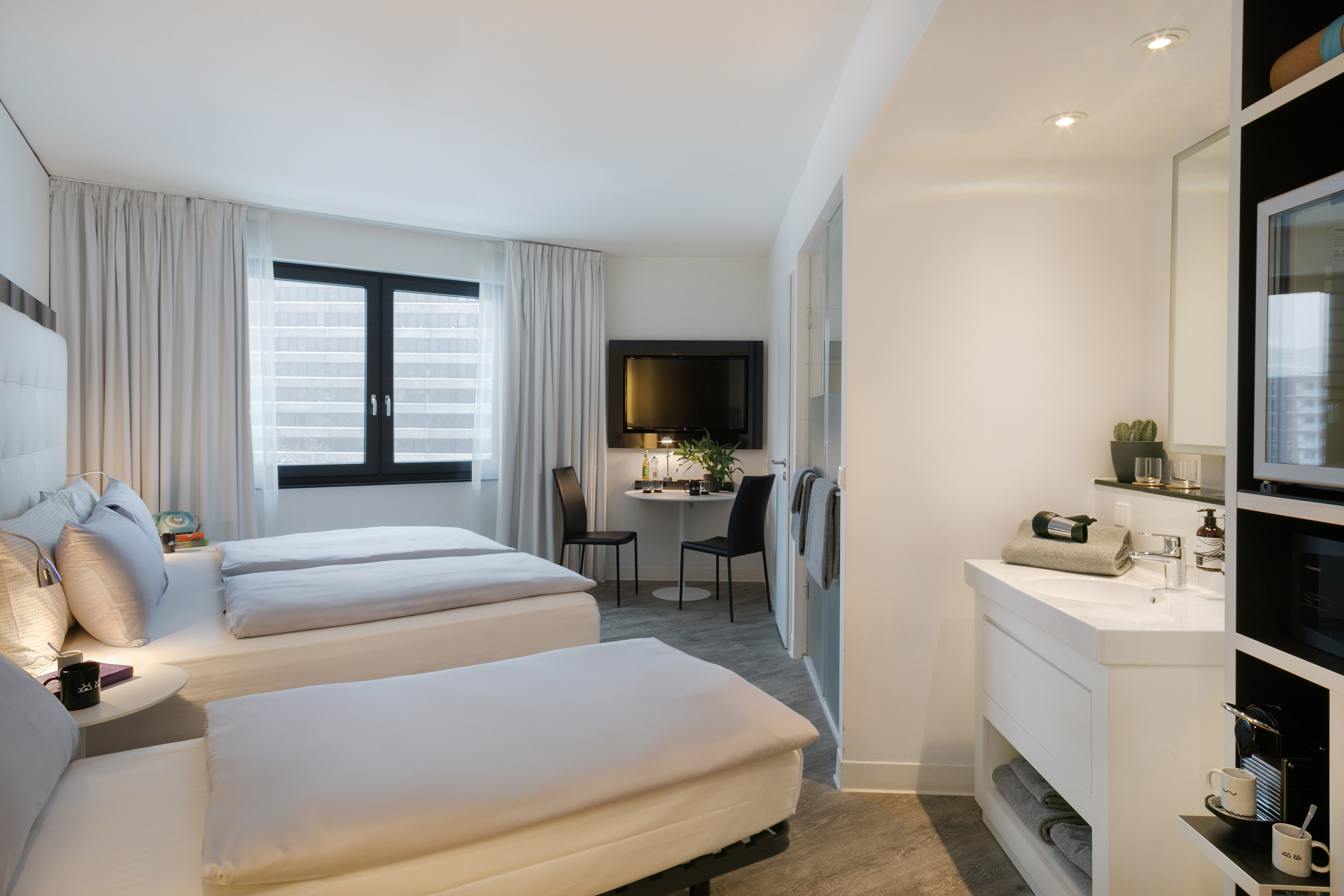 Bright, modern hotel room featuring twin beds, wall-mounted TV, and vanity.