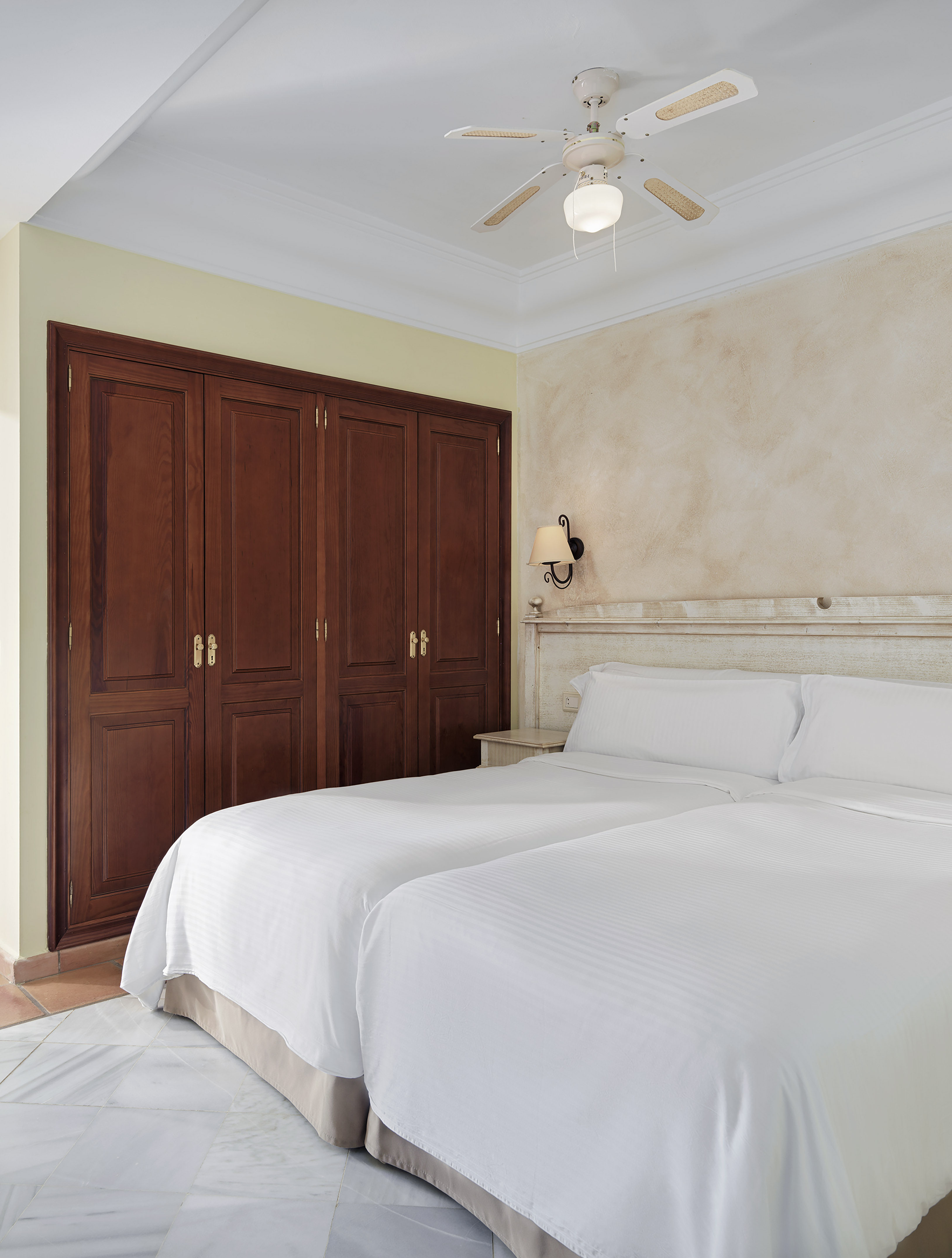 Guest room with twin beds, crisp white bedding, brown wardrobe, and ceiling fan.